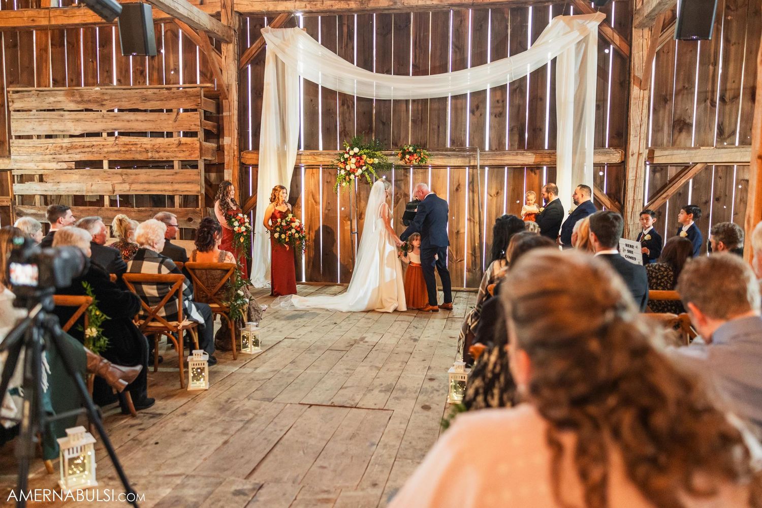 Rustic barn wedding ceremony with wooden benches and white fabric arch draped with florals and string lights.