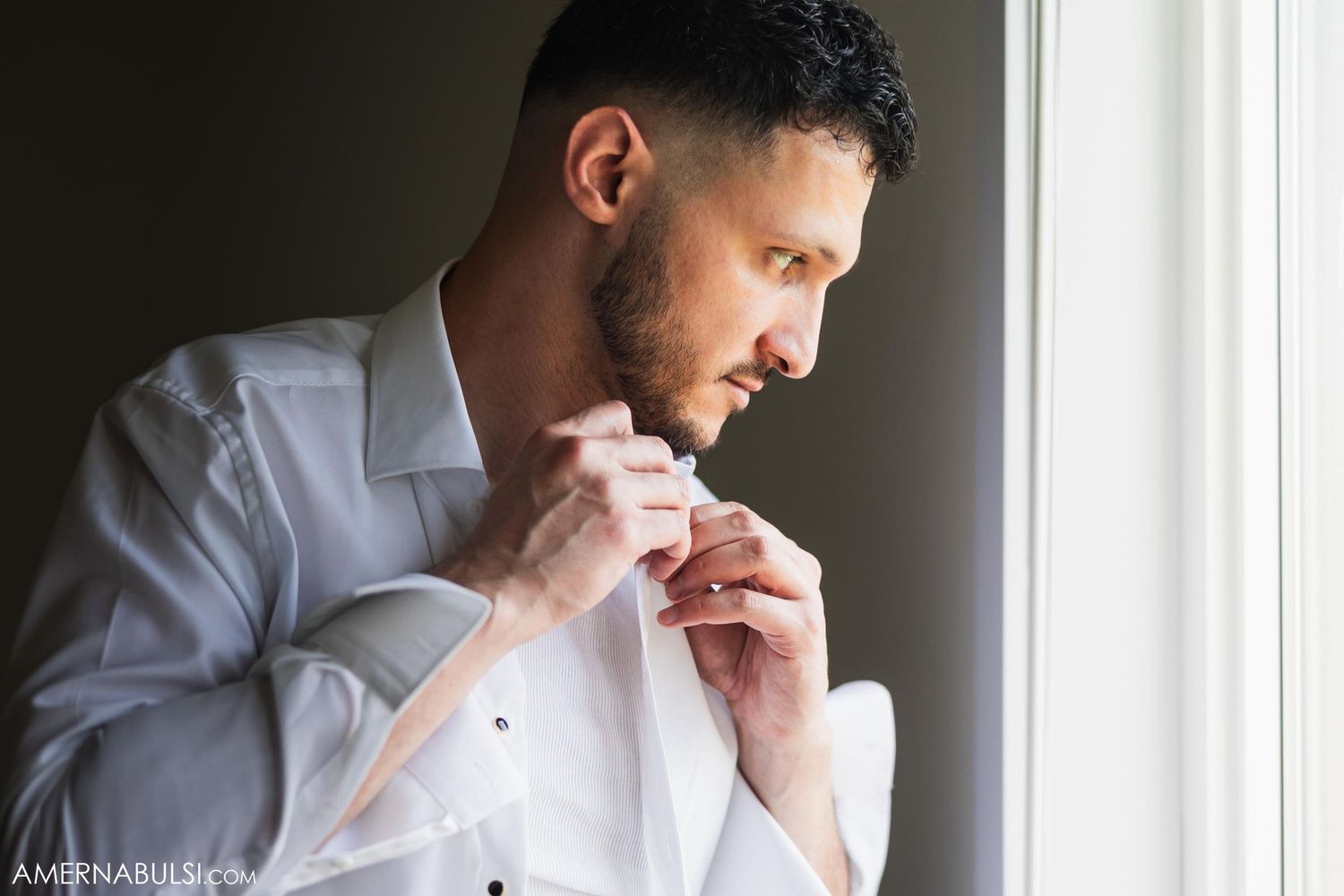 Person in white dress shirt adjusting their collar by a window.