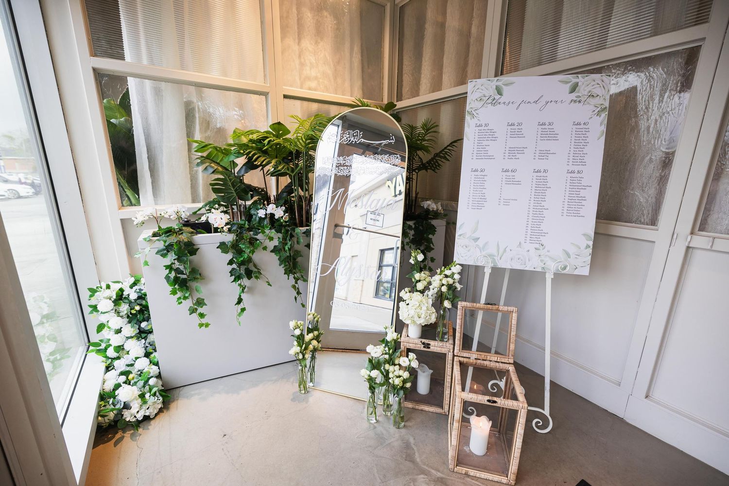 Elegant wedding seating chart display with mirror, lantern and greenery decorations.