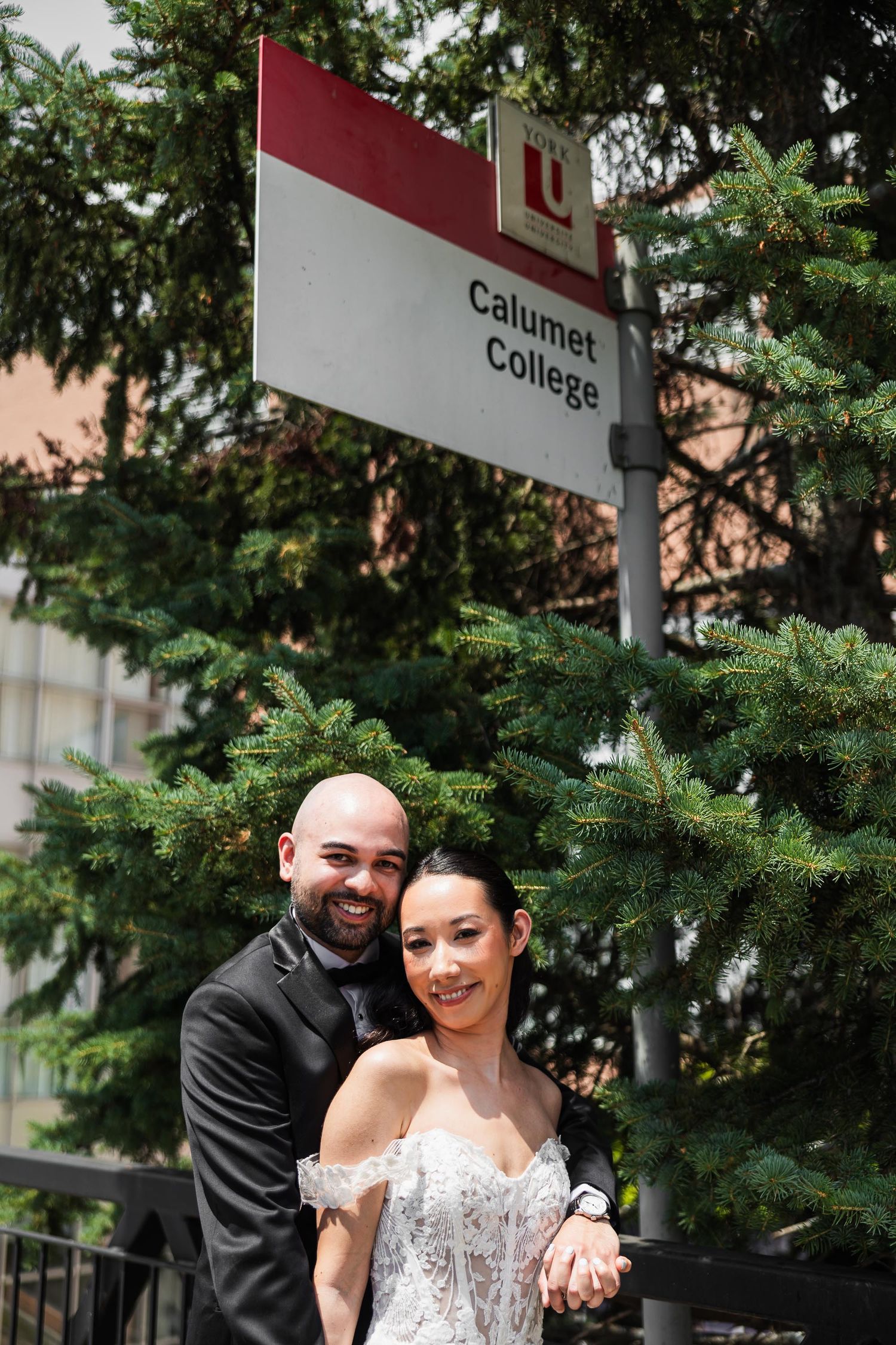 Wedding couple poses under Calumet College sign surrounded by evergreen trees.