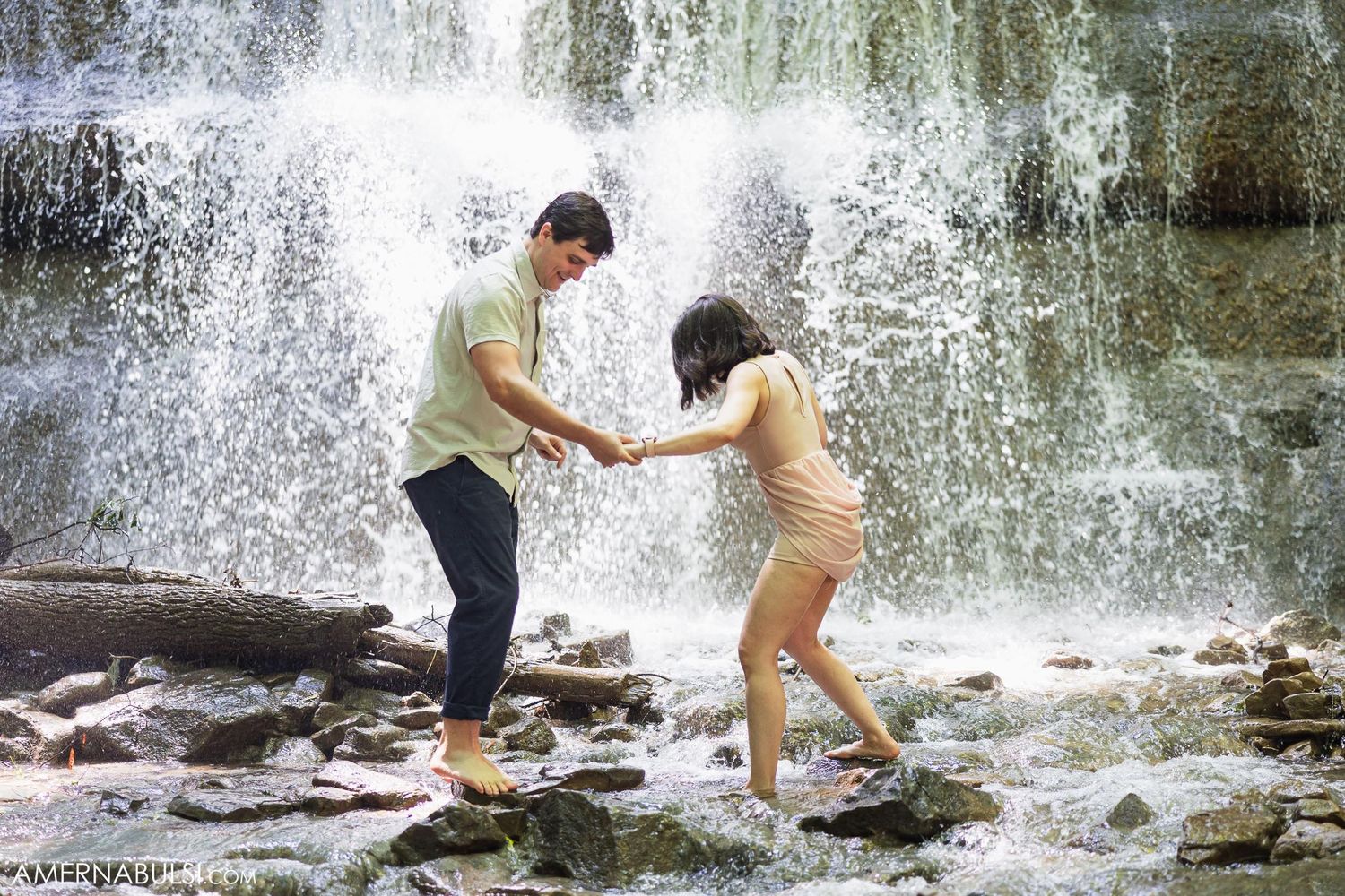 A couple holds hands while carefully navigating wet rocks in front of a misty waterfall.