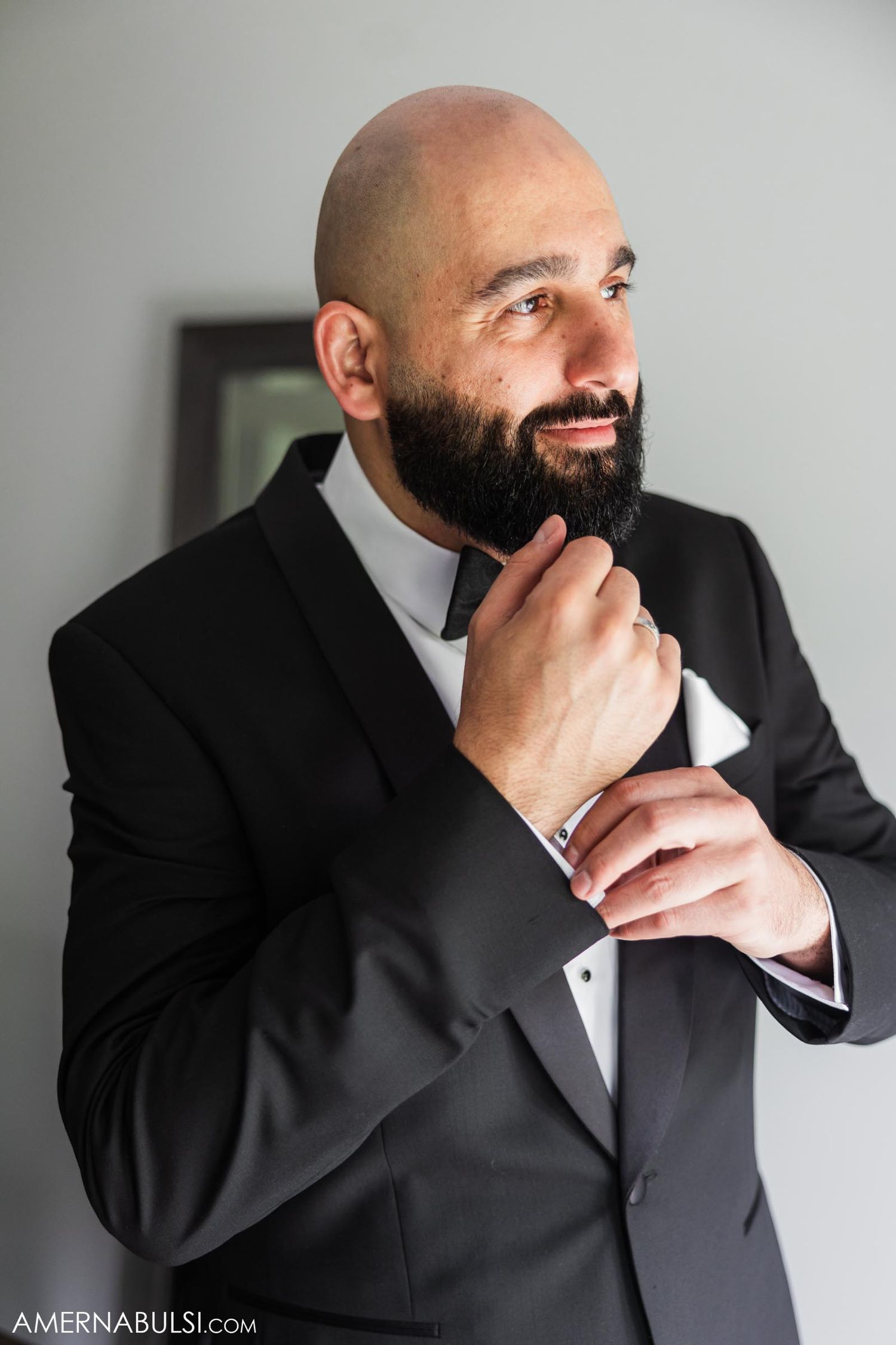 Person in black tuxedo thoughtfully touching beard while looking to the side in formal portrait.