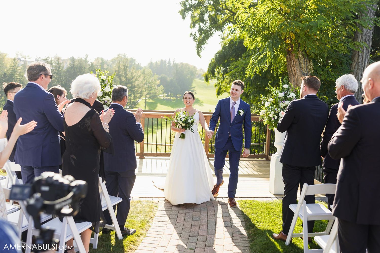 Wedding guests watching ceremony from white chairs on outdoor deck with scenic views.