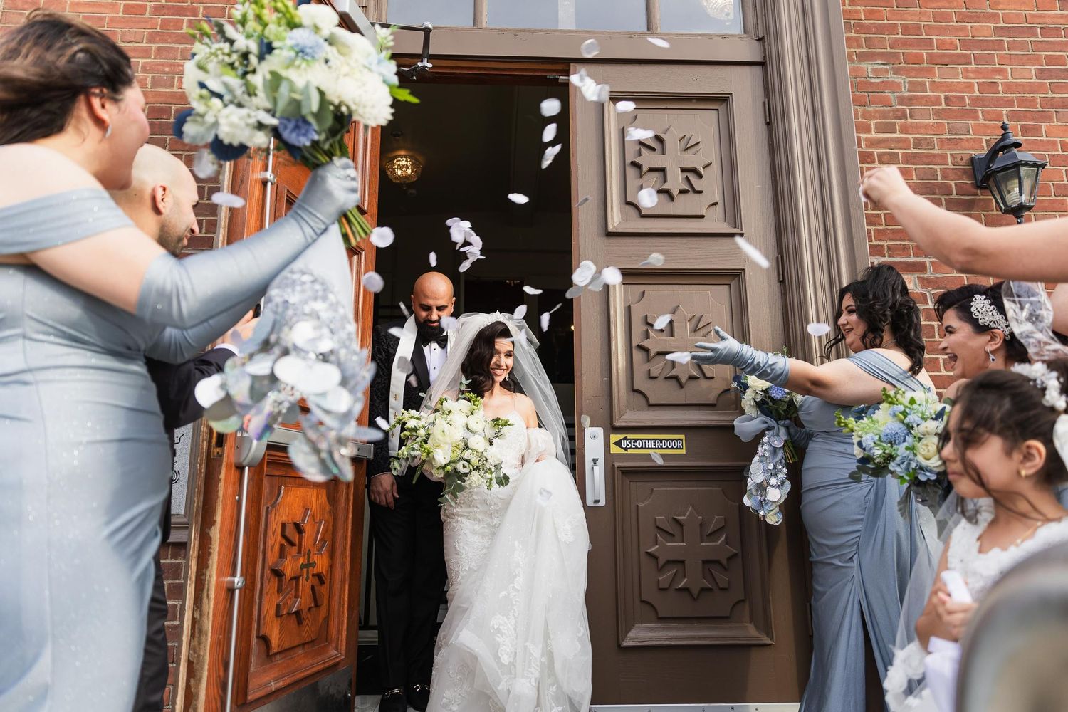 Wedding guests throw white flower petals as newlyweds exit through ornate wooden doors.