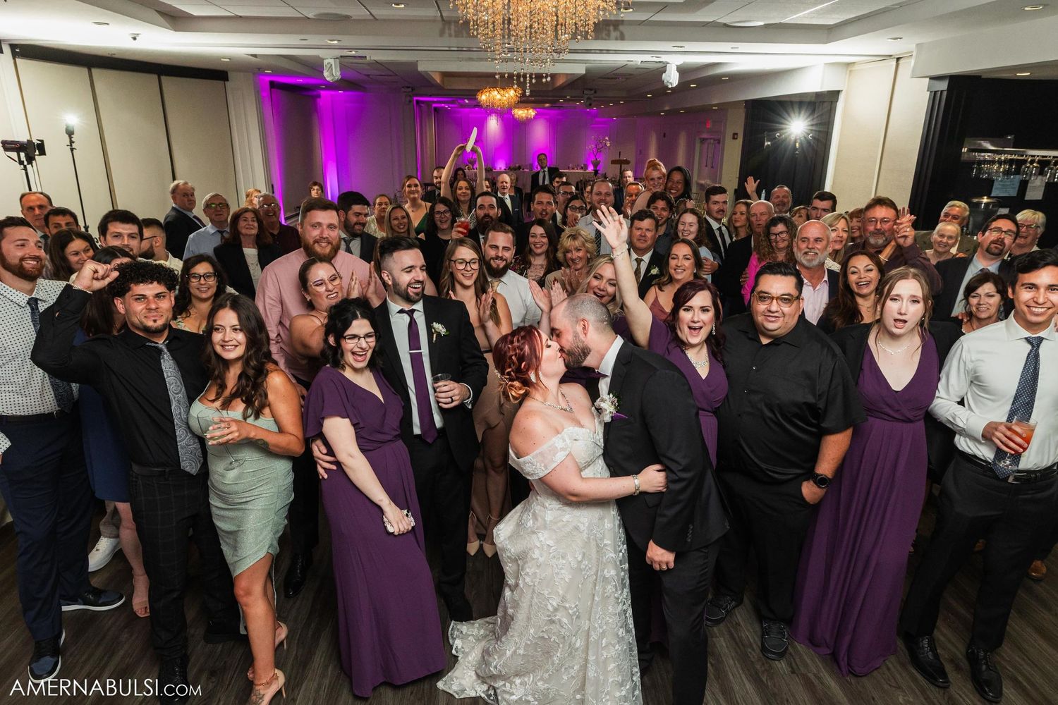 Wedding reception guests gathered in a ballroom with purple uplighting and chandelier overhead.