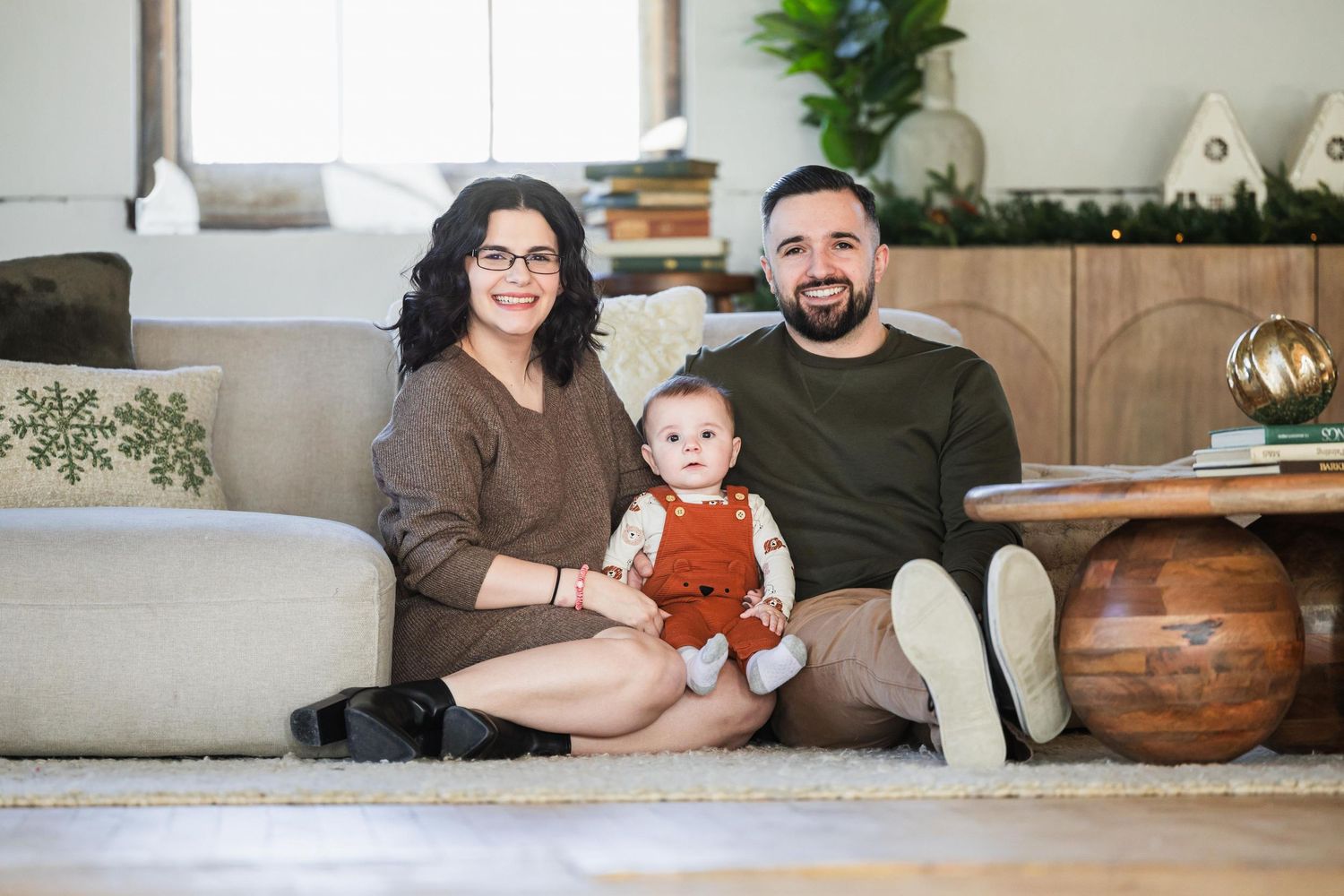 Family sitting together on couch with baby in orange outfit and decorative pumpkins nearby.