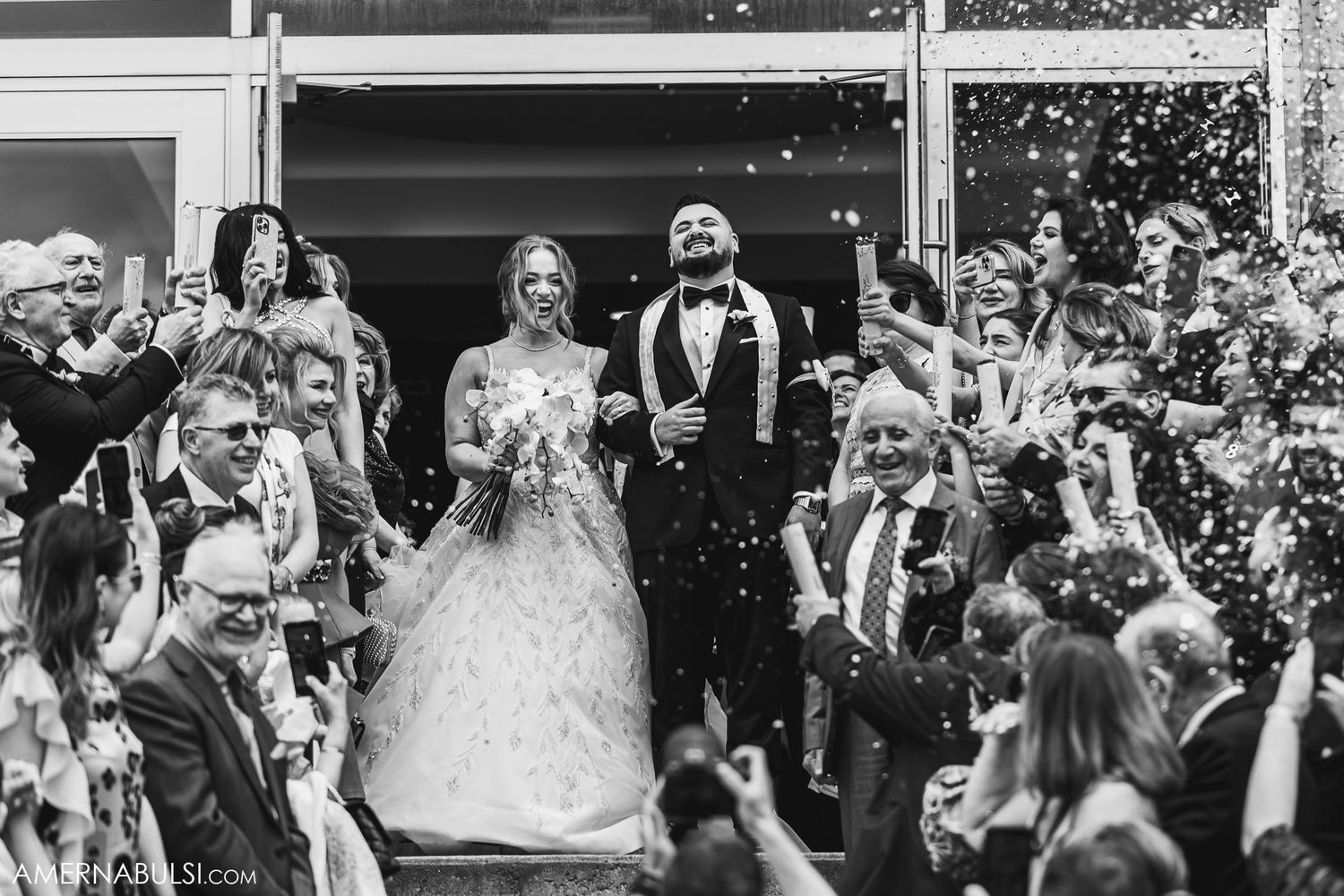 Black and white capture of wedding celebration with confetti shower and cheering crowd.