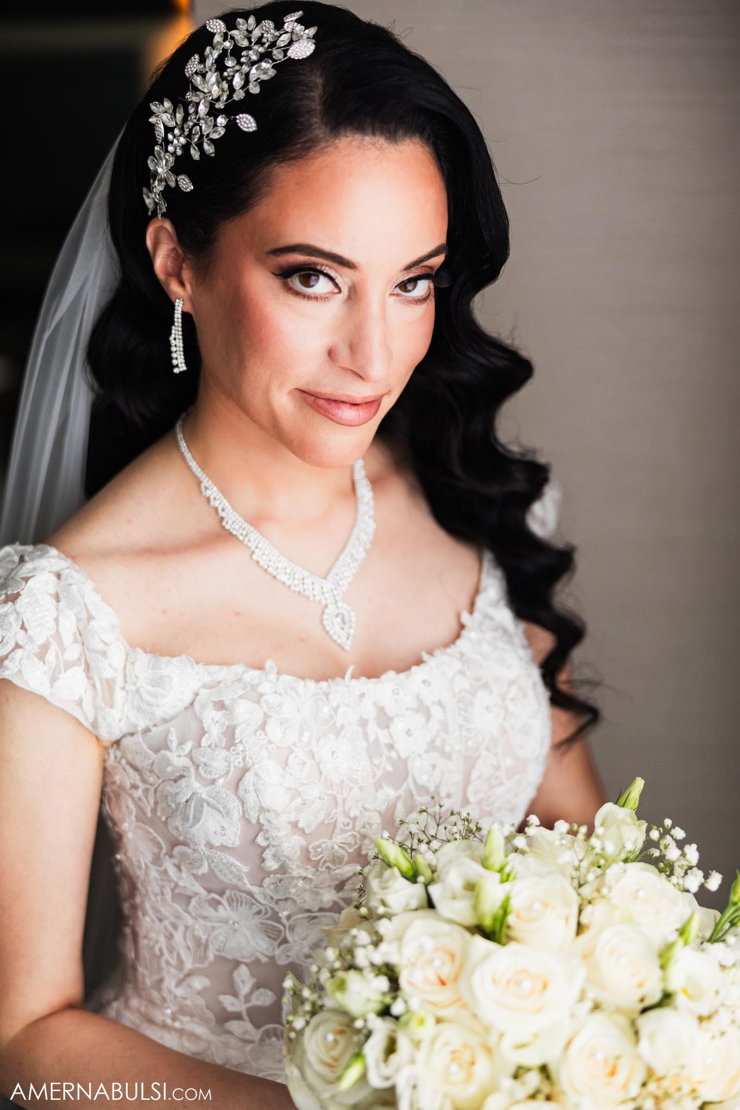 Elegant bride in lace dress with crystal accessories holds white rose bouquet.