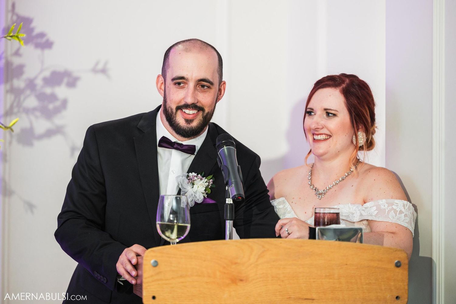 Couple in formal attire smiling while standing at wooden podium during wedding reception.