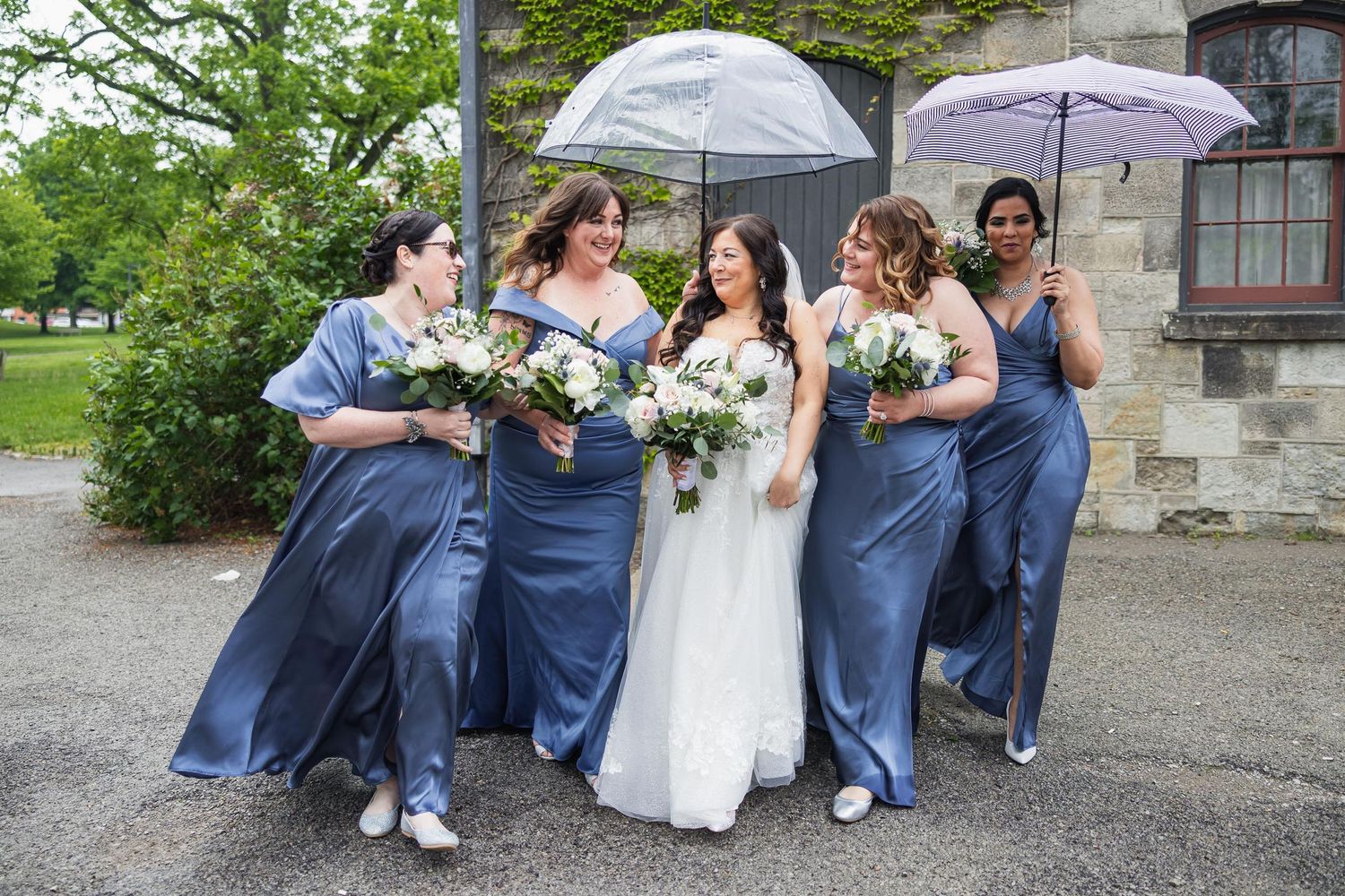 Bridal party holding umbrellas poses together in blue dresses with white bouquets.