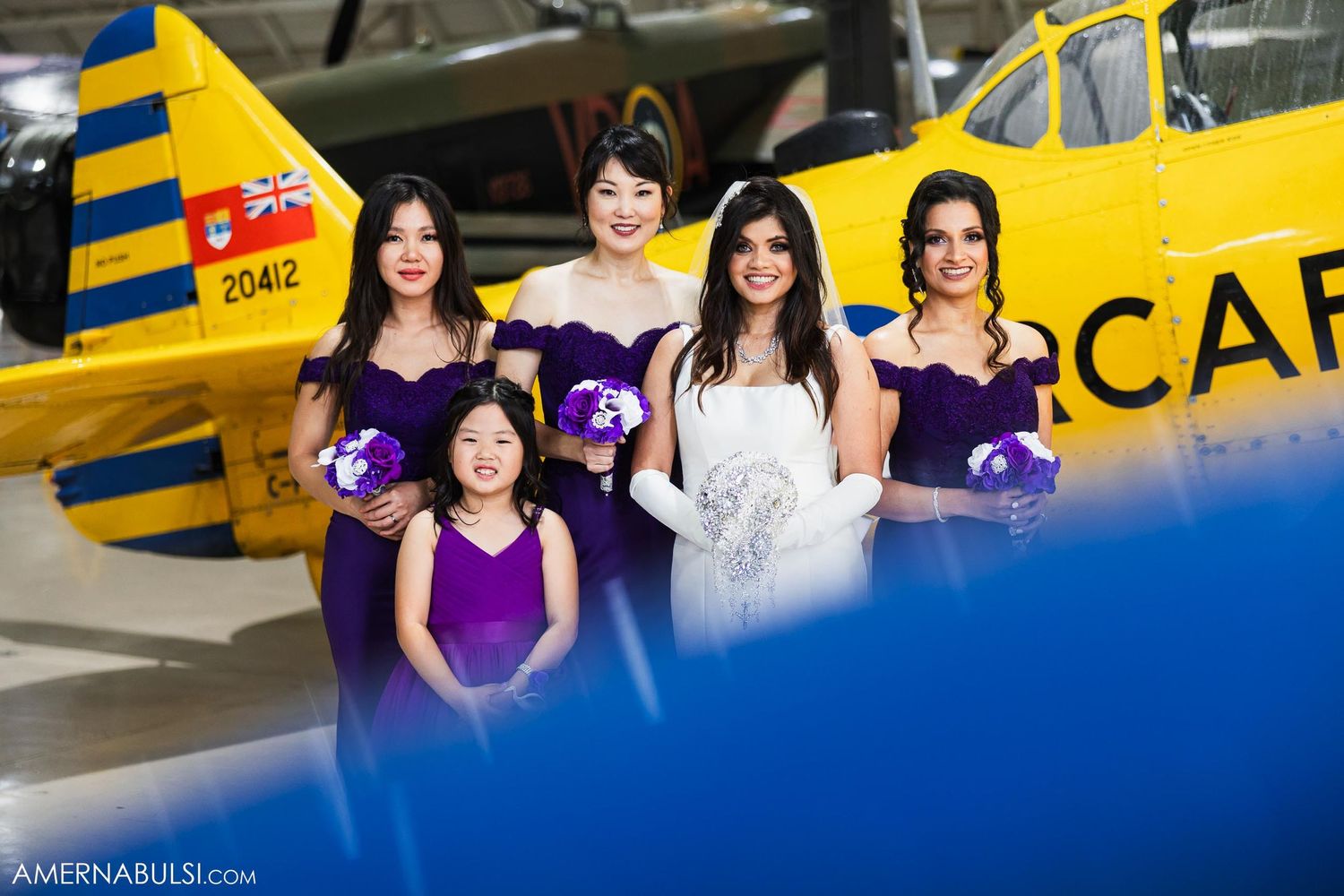 A bridal party in purple dresses poses with a yellow vintage aircraft in the background.