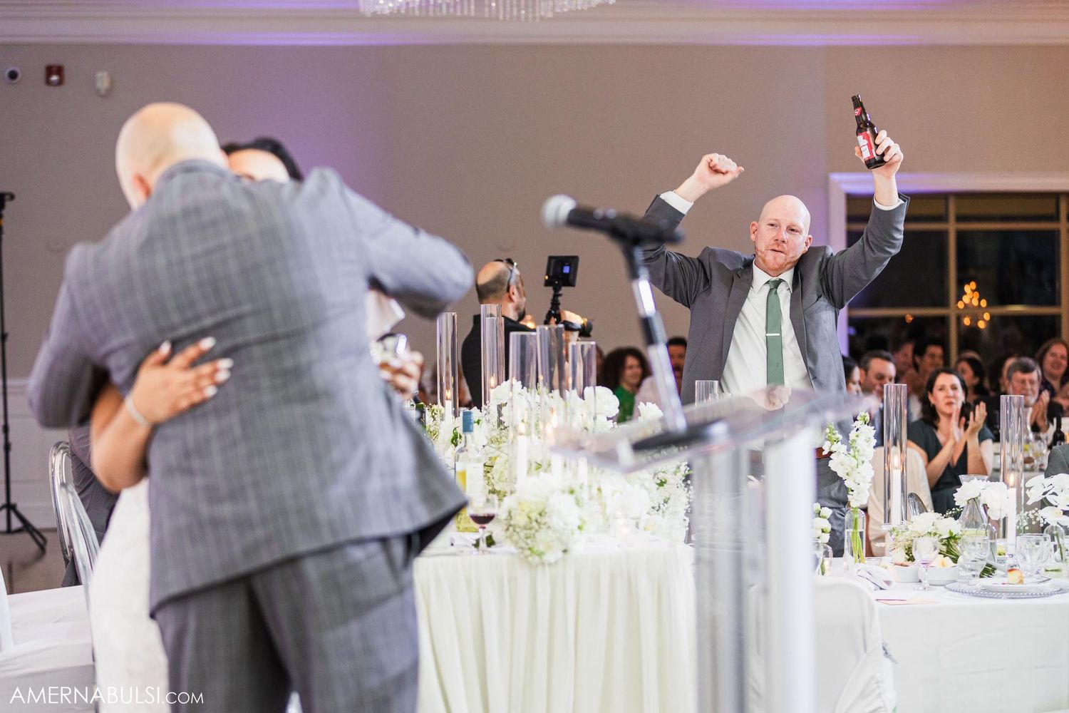 Wedding guests celebrating with raised arms at a reception dinner with elegant white table settings.