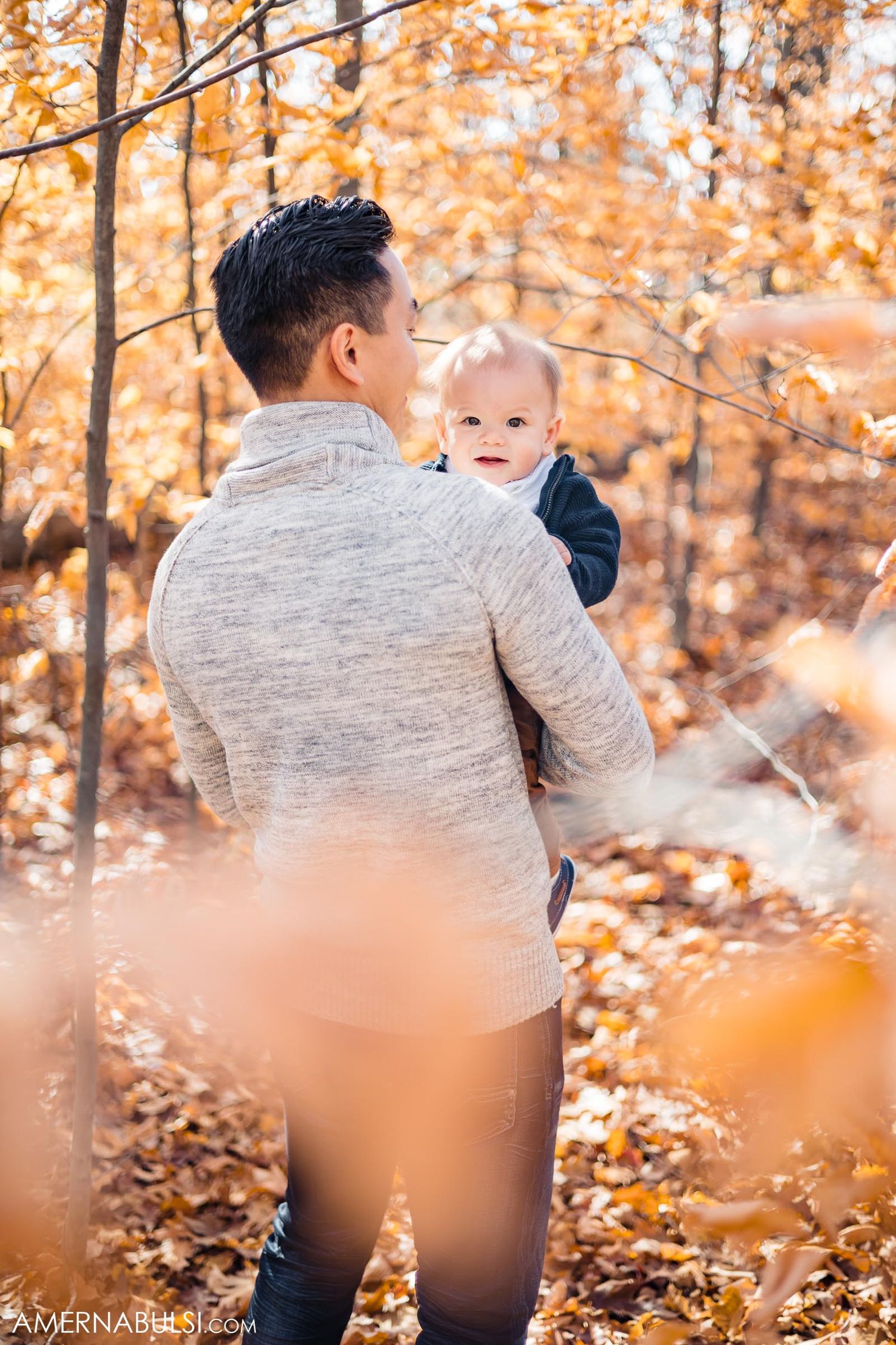 Parent holding infant while standing among fallen autumn leaves in forest.