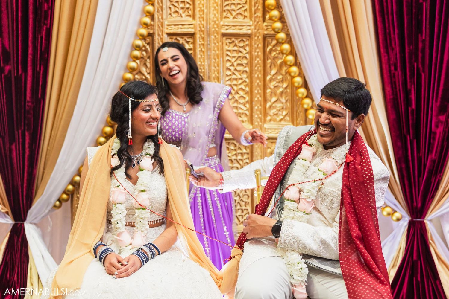 Wedding party shares lighthearted moment during Indian ceremony with decorative drapery and backdrop.