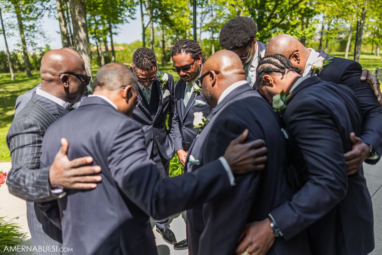 Group of men in dark suits gather in emotional huddle outdoors.
