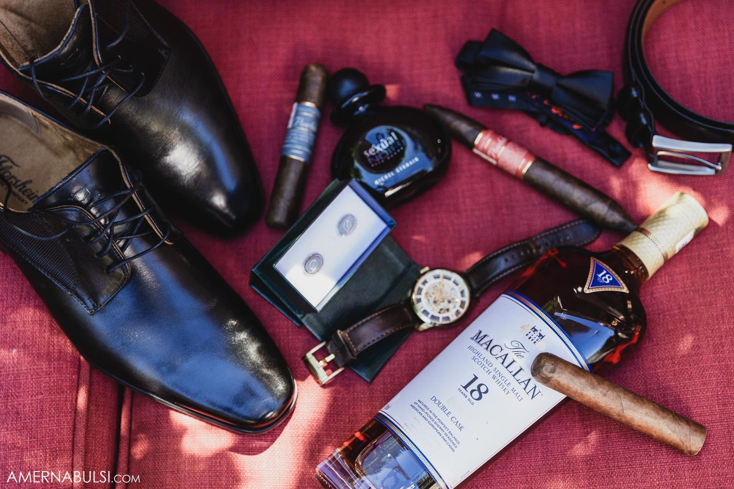 Wedding day accessories including formal shoes, cigar, cologne, and watch arranged on red fabric.