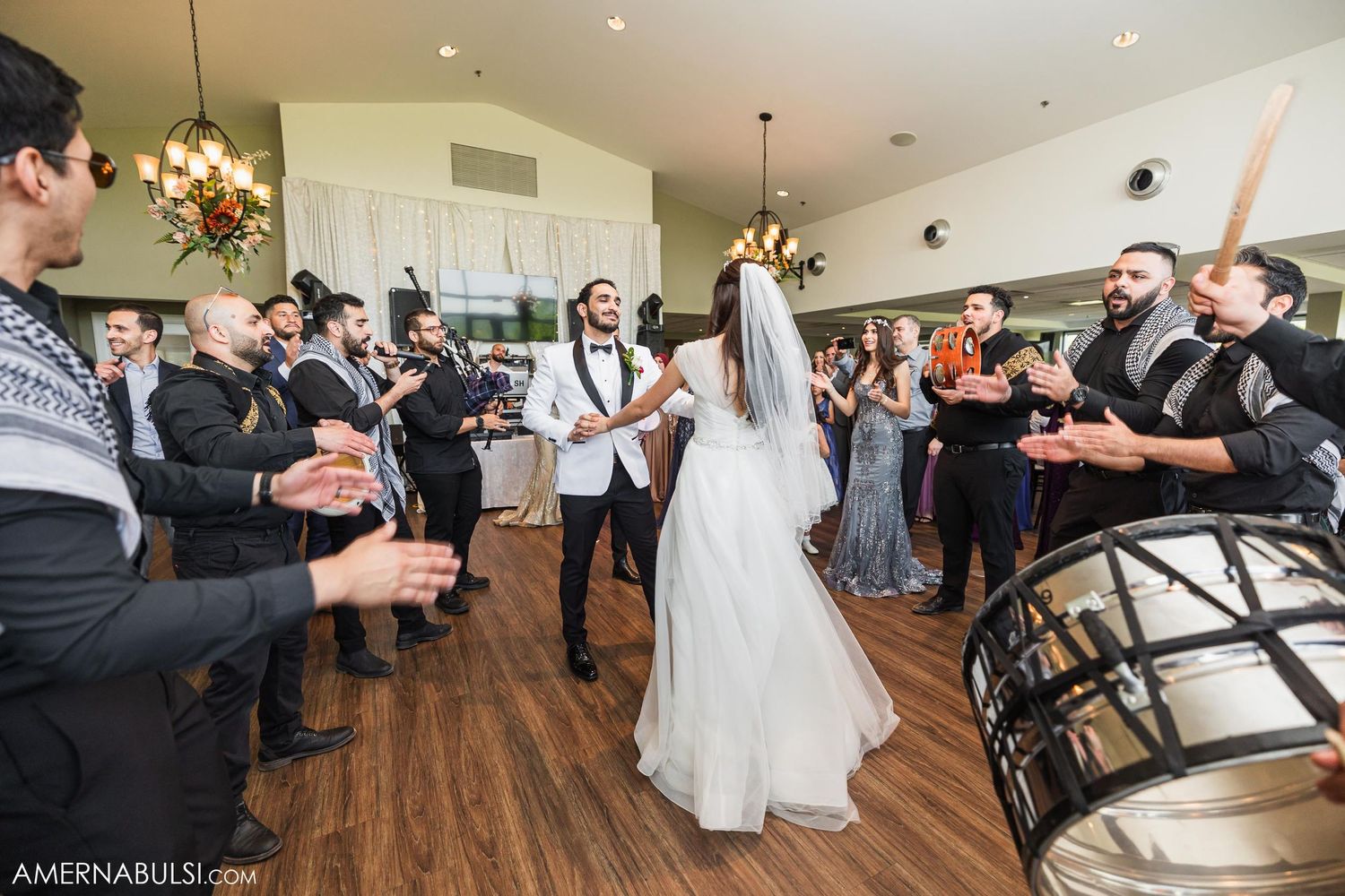 Wedding guests clap and celebrate as newlyweds dance in a reception hall with hardwood floors.
