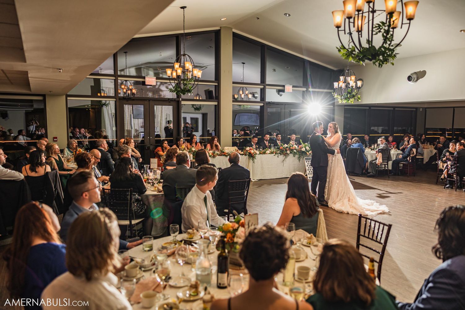 Wedding celebration in an elegant reception hall with chandeliers as couple dances surrounded by seated dinner guests.