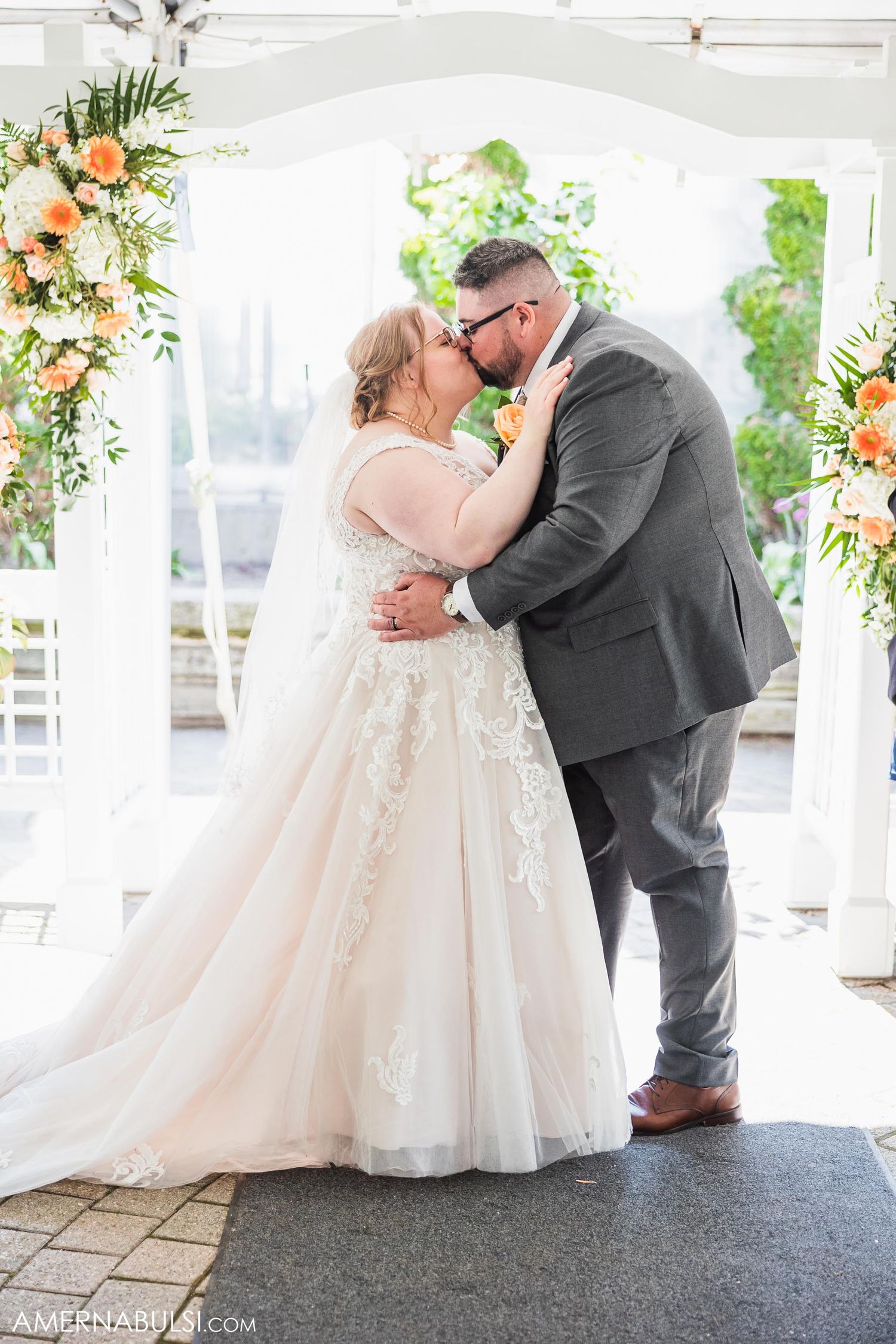 Couple sharing romantic kiss under floral-decorated wedding arch during ceremony.