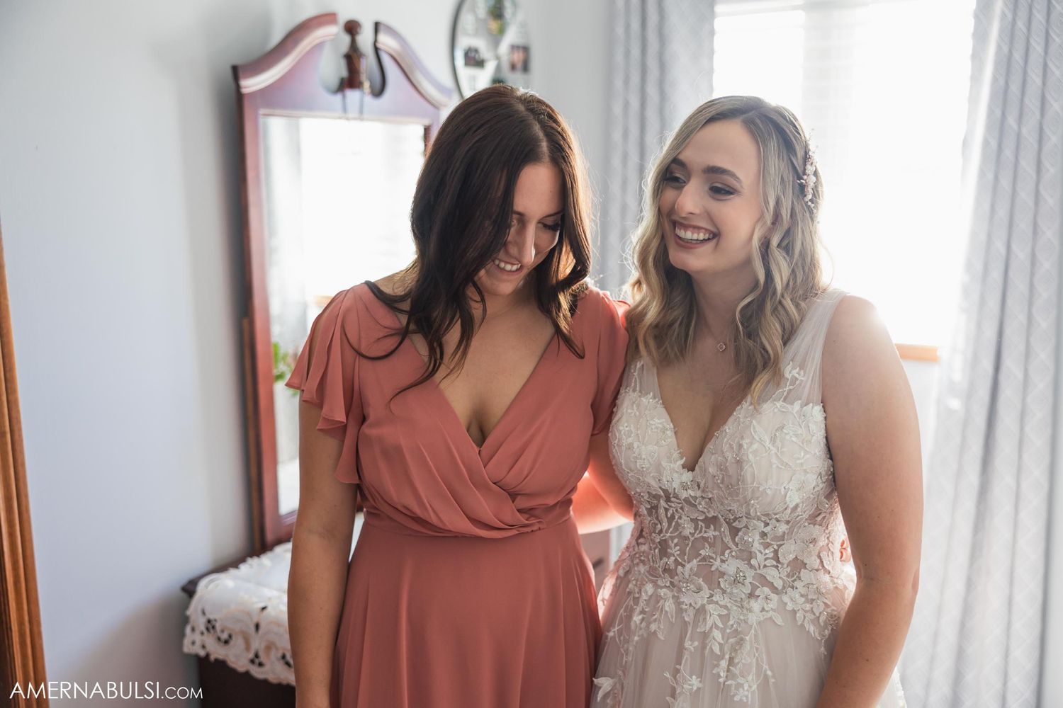 Two women share a joyful moment in a bedroom, one wearing coral and one in a white lace wedding dress.