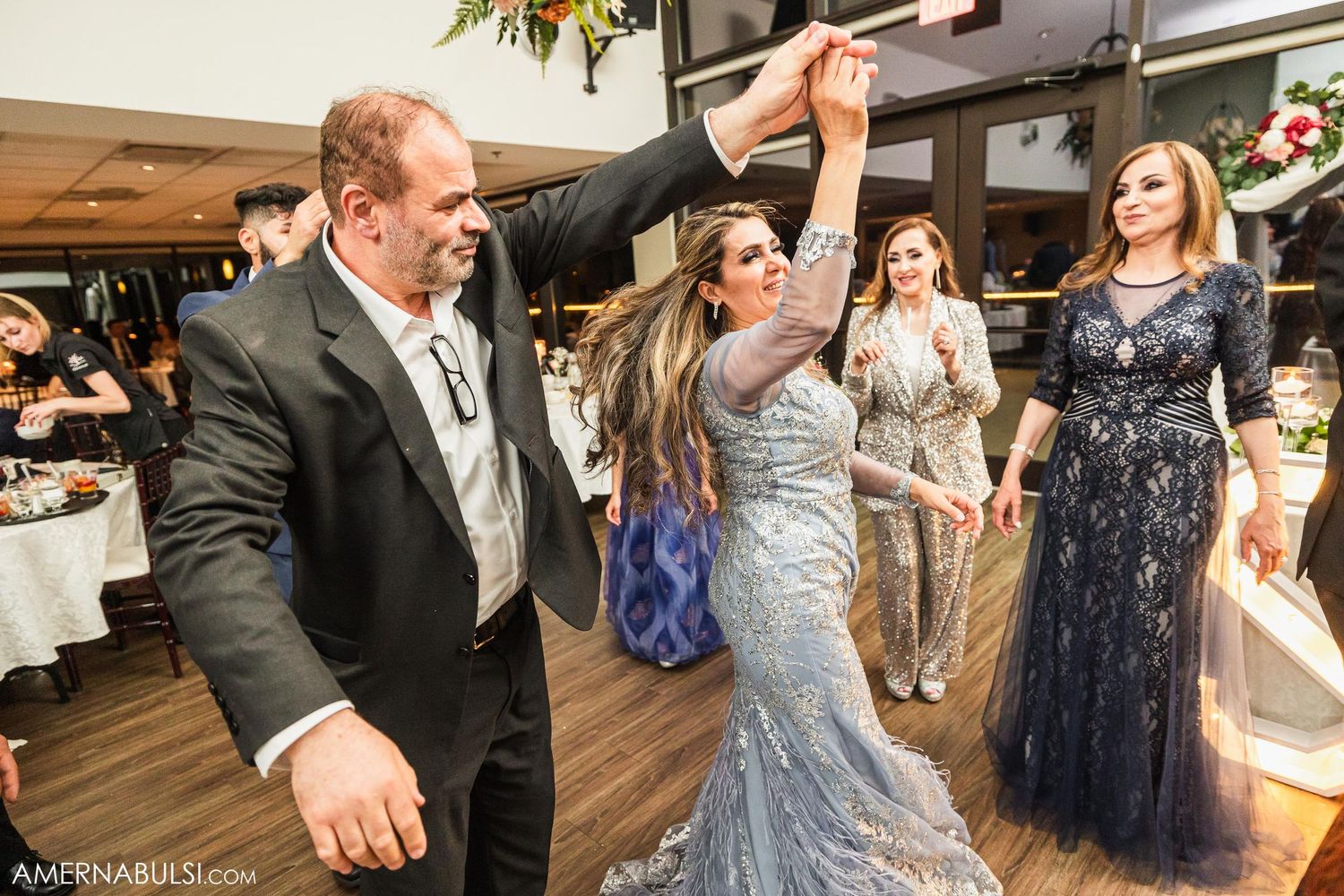Wedding guests dancing enthusiastically on a wooden dance floor at an indoor reception venue.