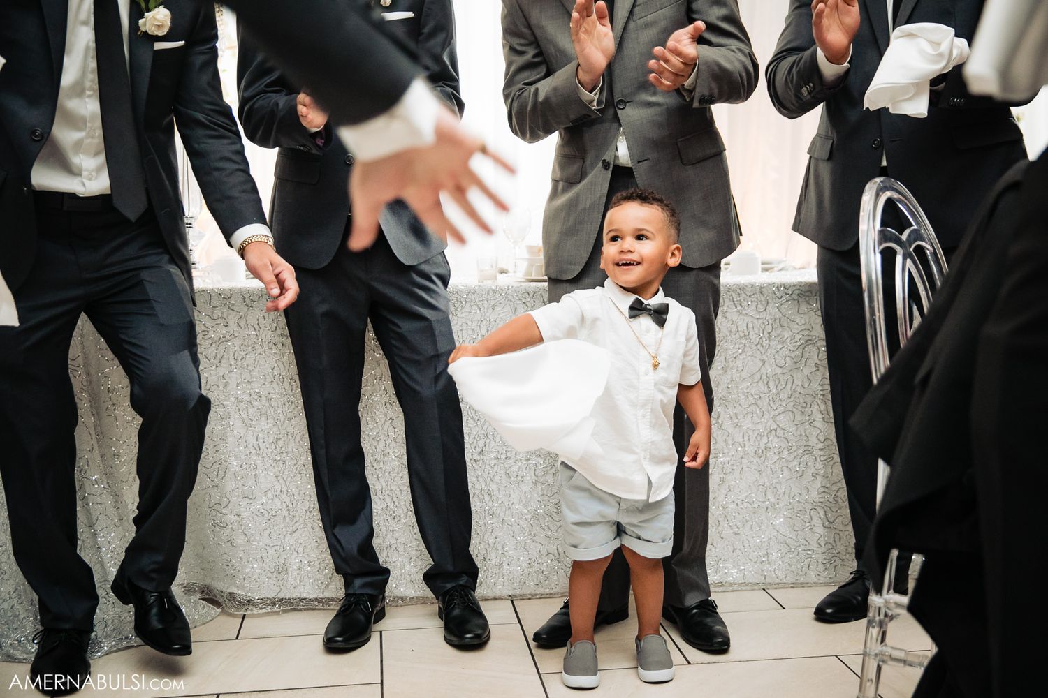 Young ring bearer in white suit dances among groomsmen at wedding celebration.