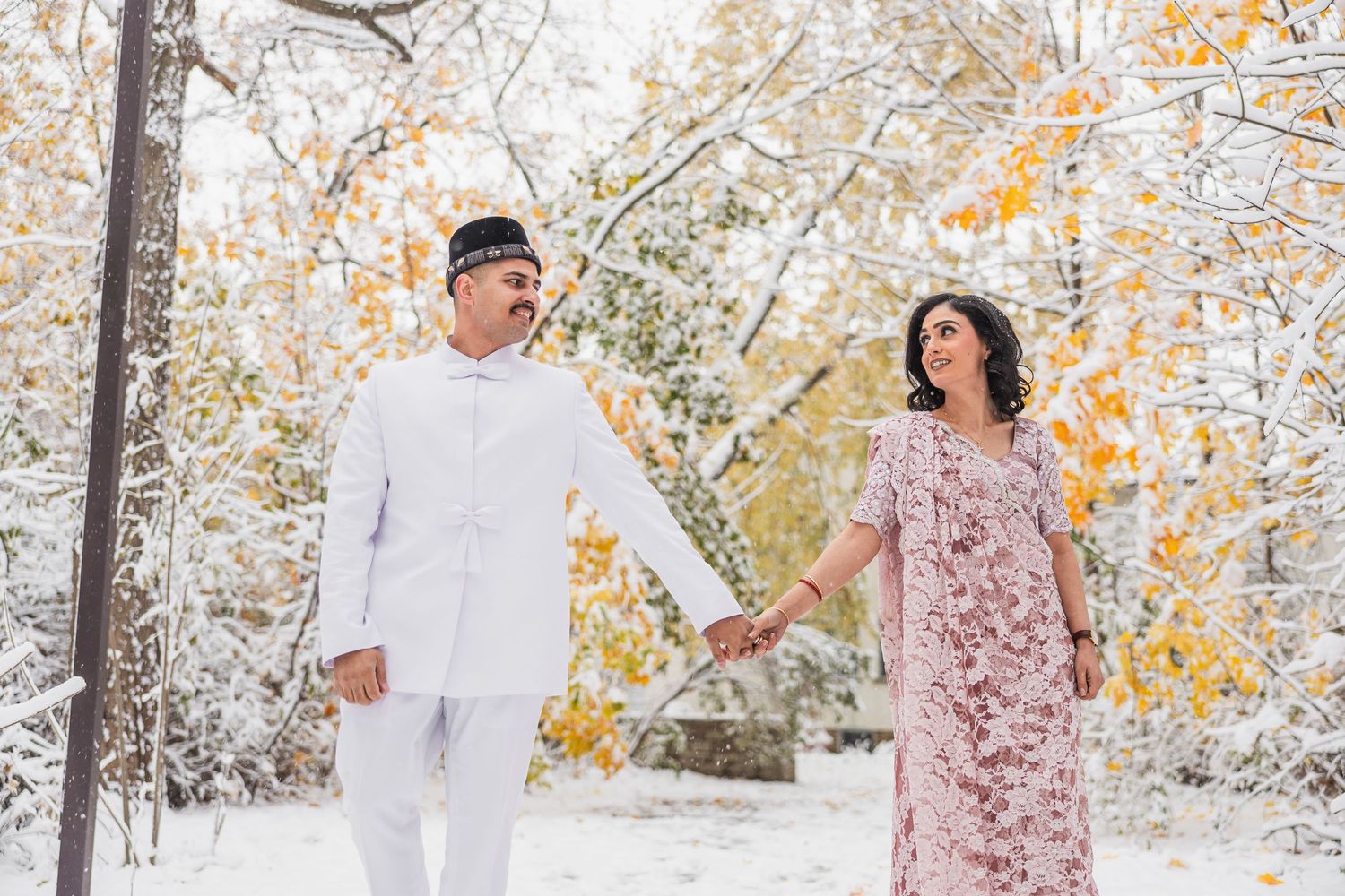 Couple in white and pink outfits hold hands while walking in a snowy autumn forest setting.