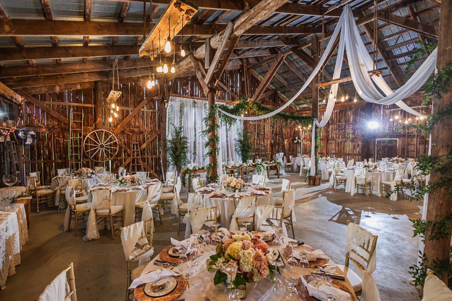 Rustic barn wedding reception venue decorated with twinkling lights and white fabric draping along wooden beams.