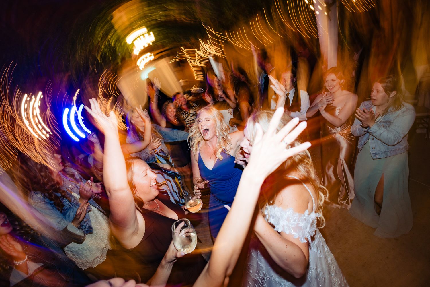 Wedding reception guests dancing and celebrating on a dimly lit dance floor under colorful lighting.
