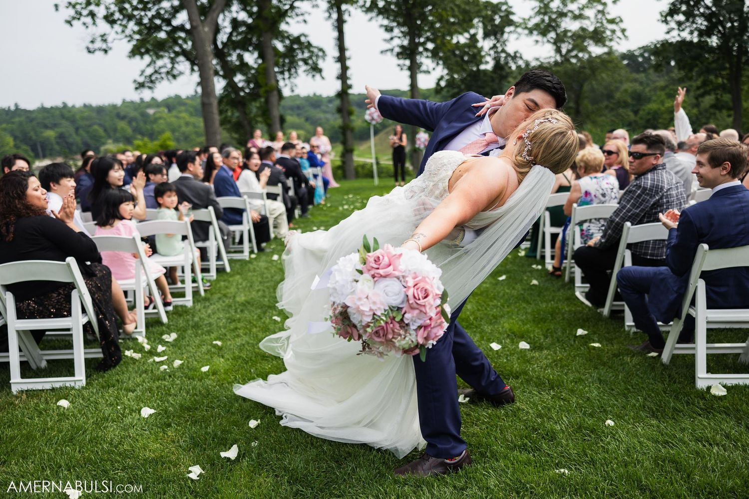 Romantic dip kiss captured at outdoor wedding ceremony with guests seated on white chairs.