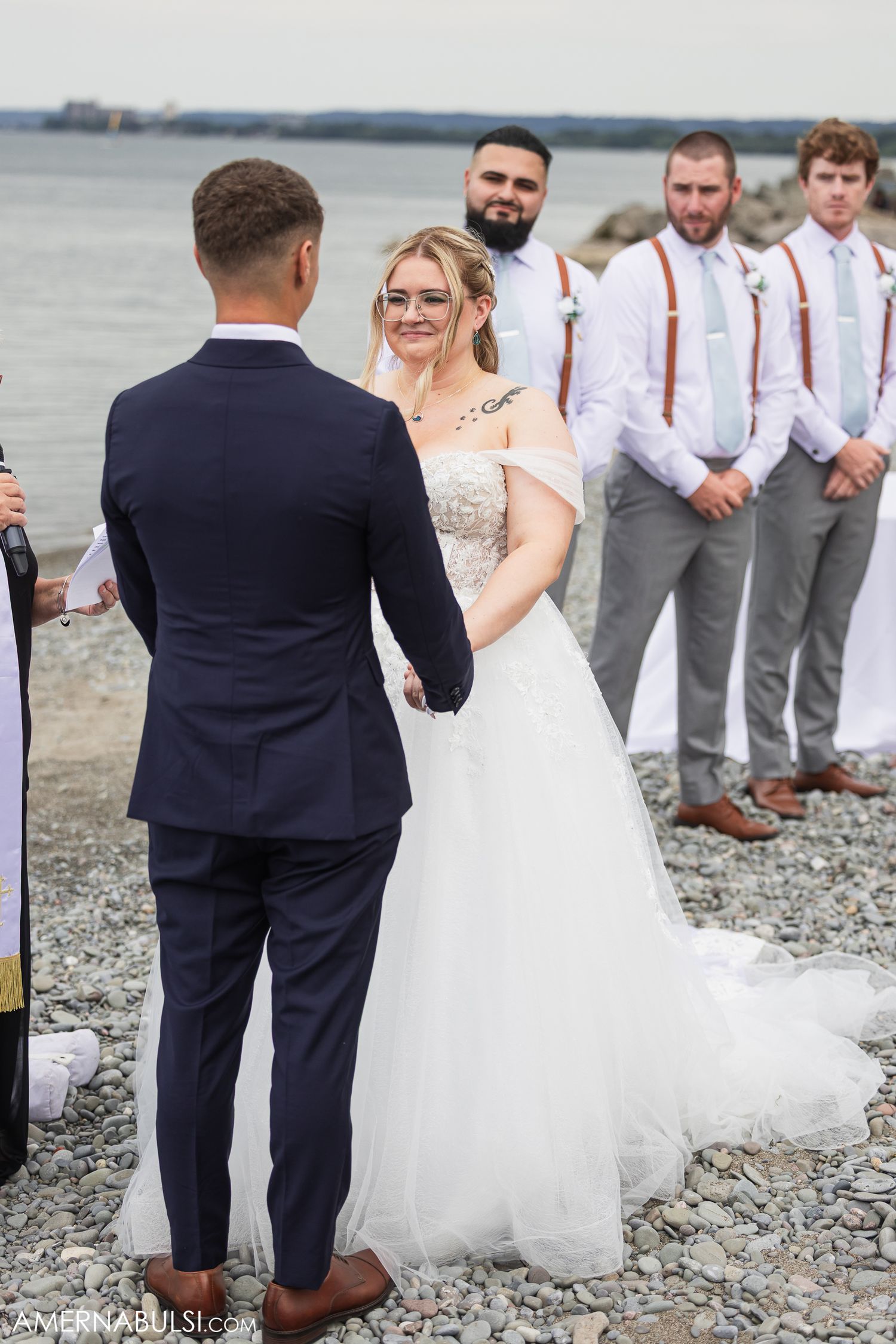 Wedding party gathered on a rocky beach shore with bridesmaids in light dresses and groomsmen in gray suits.