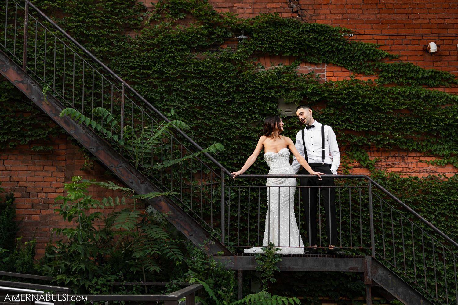 Couple in wedding attire pose on metal staircase against brick and ivy wall.