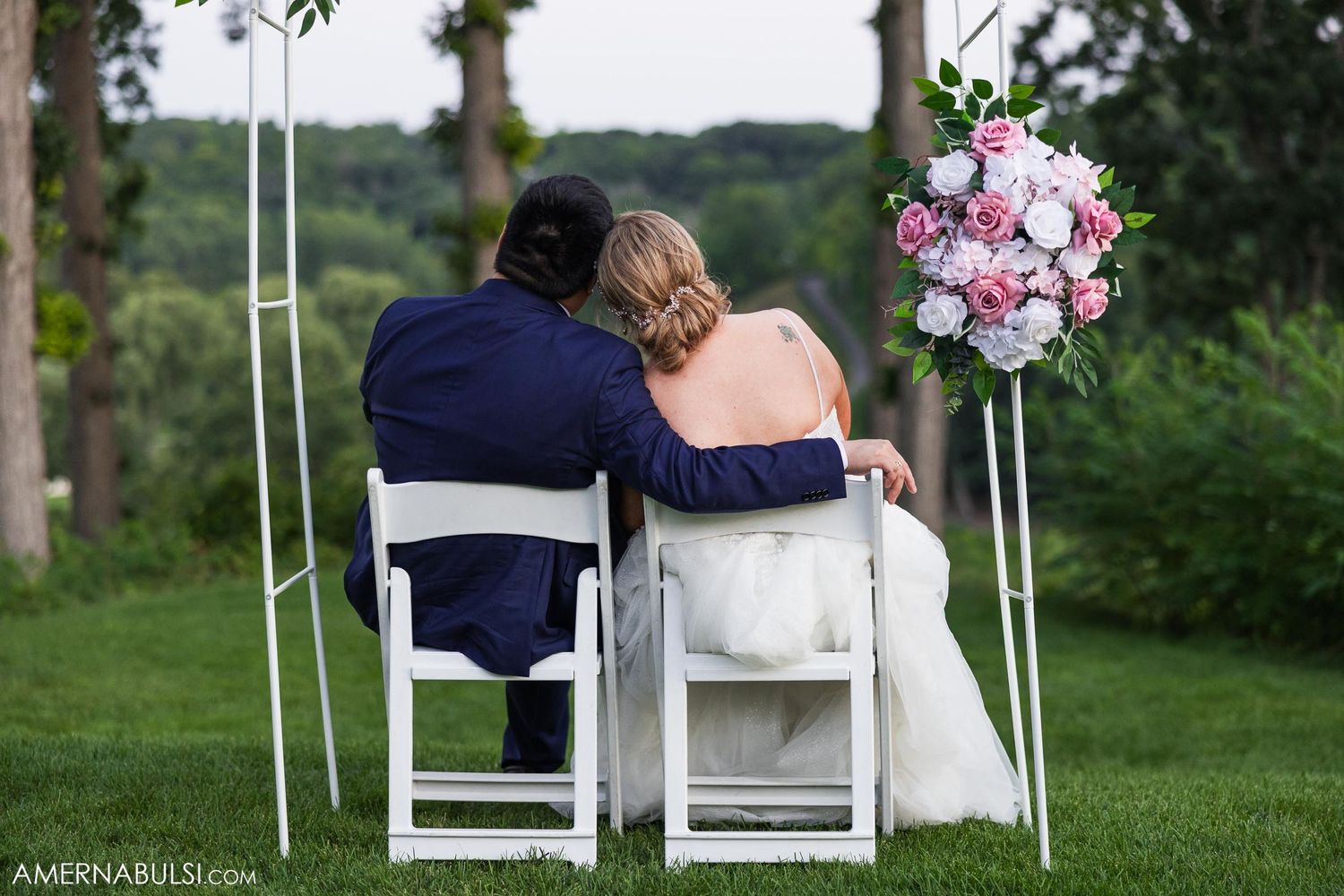 Newlyweds sitting together on white chairs in a garden setting with floral arrangements.