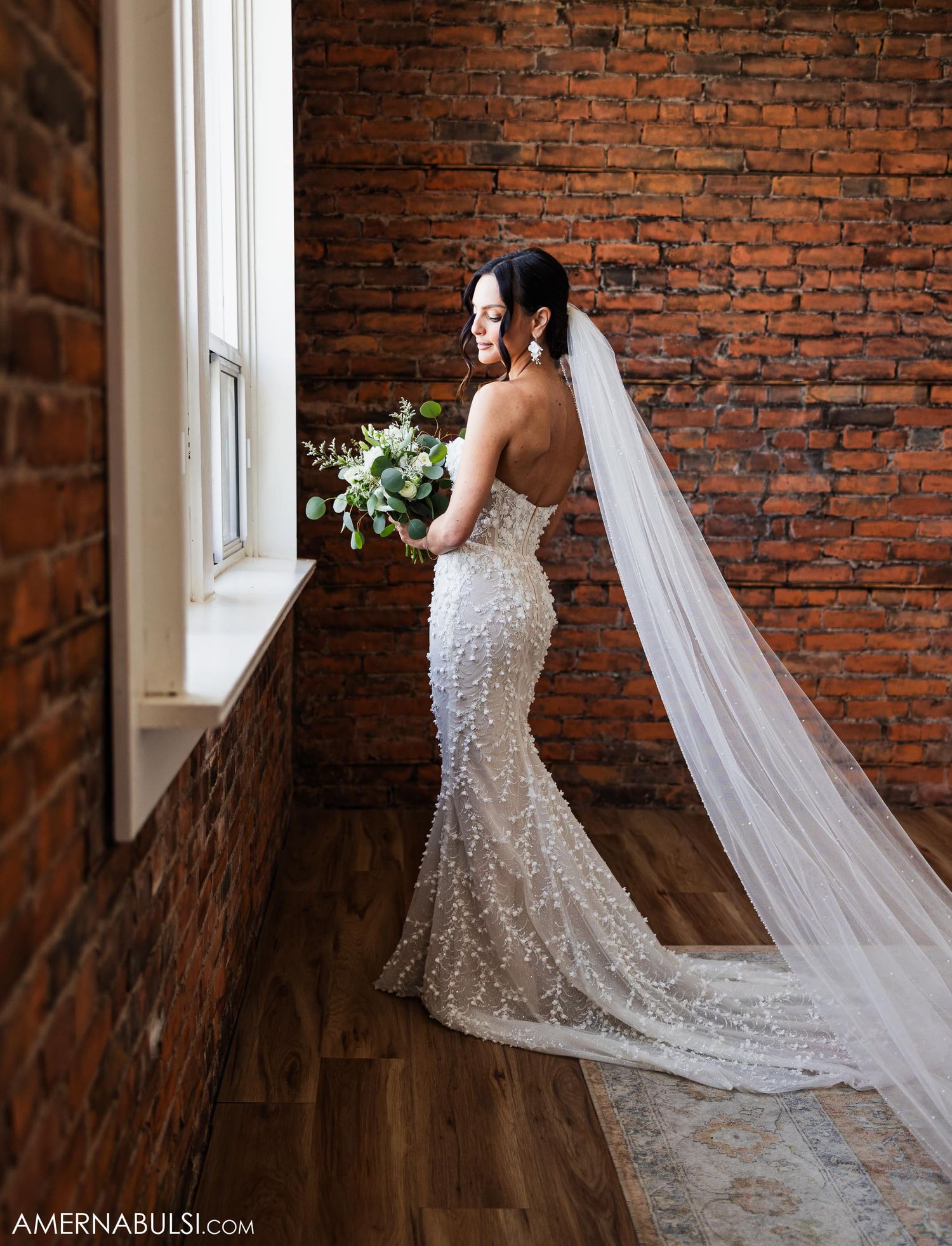 A bride in a fitted lace wedding gown with long veil poses by a brick wall near a window.