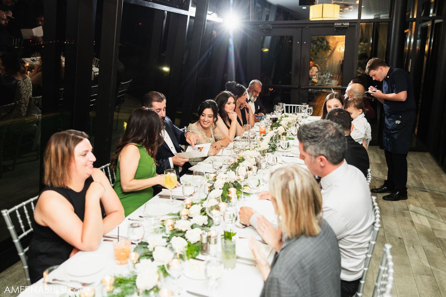 Elegant dinner party with guests seated at a long table in dimly lit restaurant setting.