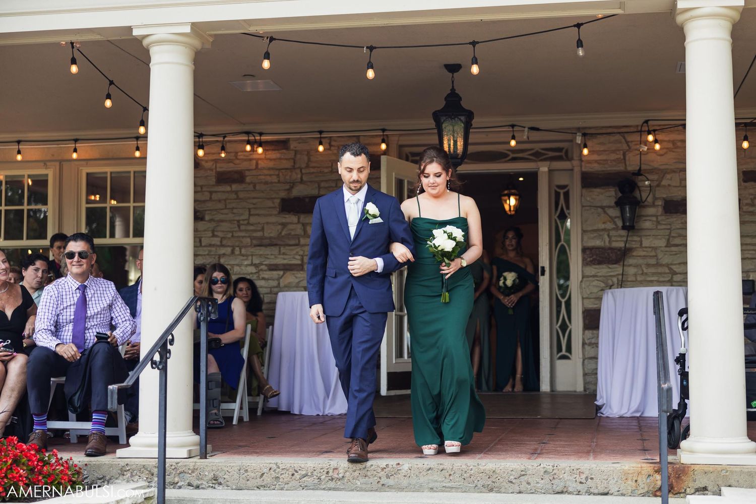 A couple walks down stone steps at an elegant venue with white columns and string lights.
