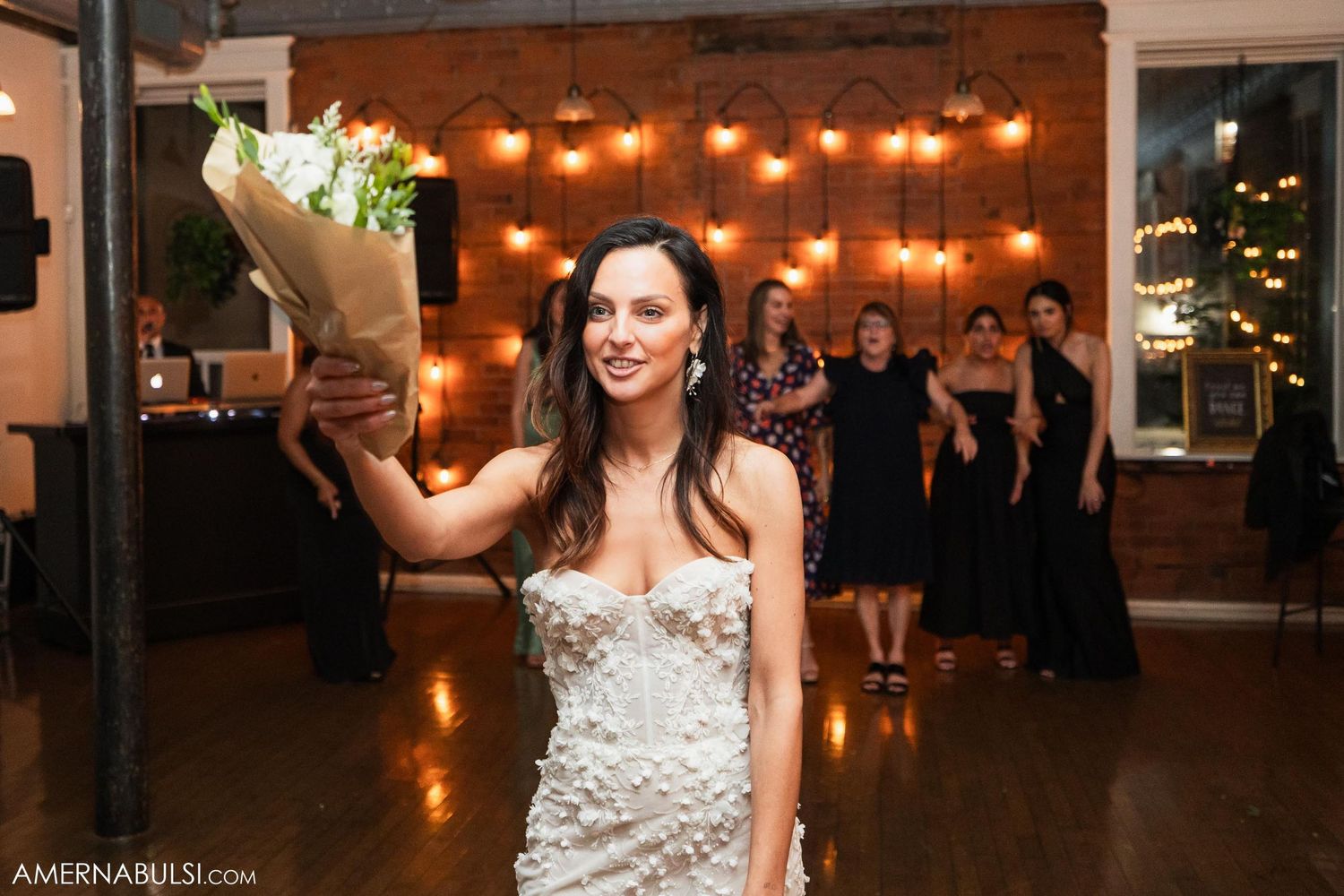 Person in white wedding dress holding bouquet in venue with string lights.