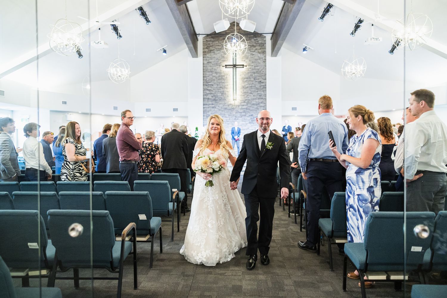 Wedding guests seated in white chairs during an elegant indoor ceremony with vaulted ceiling and crystal chandeliers.