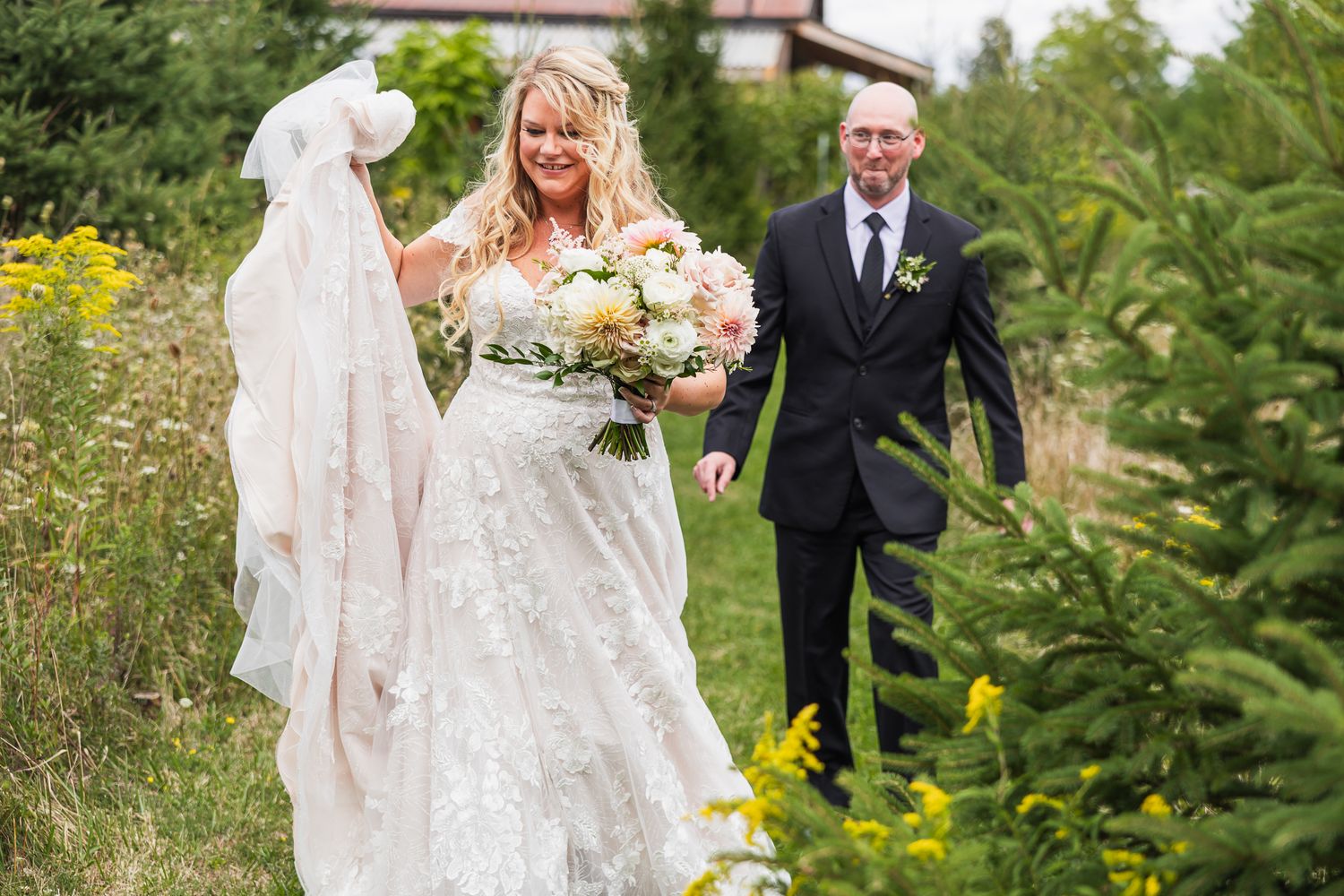 A wedding couple walks through a garden path surrounded by lush green trees and foliage.