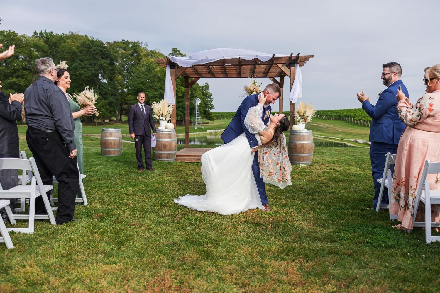 Outdoor wedding ceremony under wooden pergola with guests watching from white chairs.
