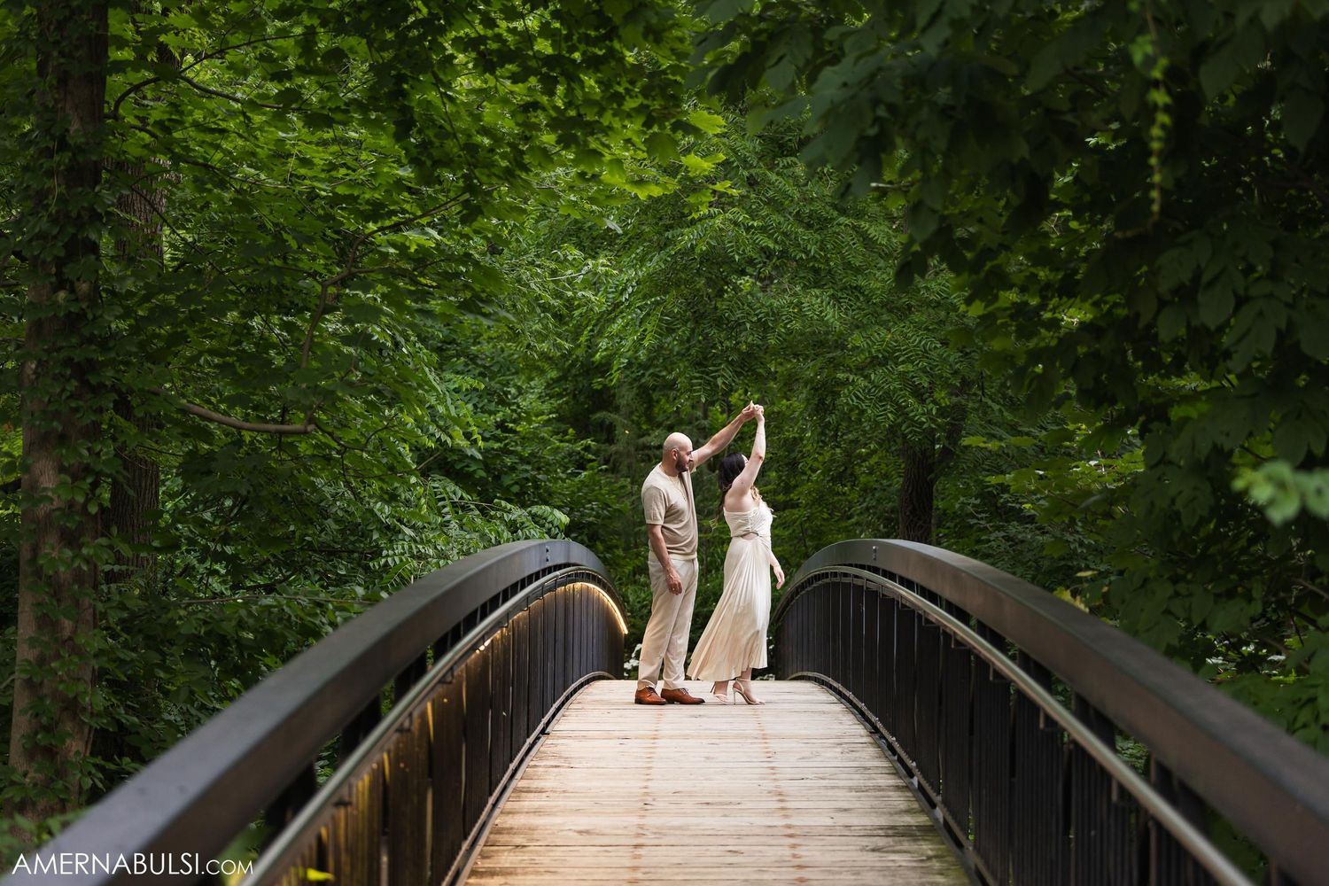 A couple celebrates on a curved wooden bridge surrounded by lush green forest.