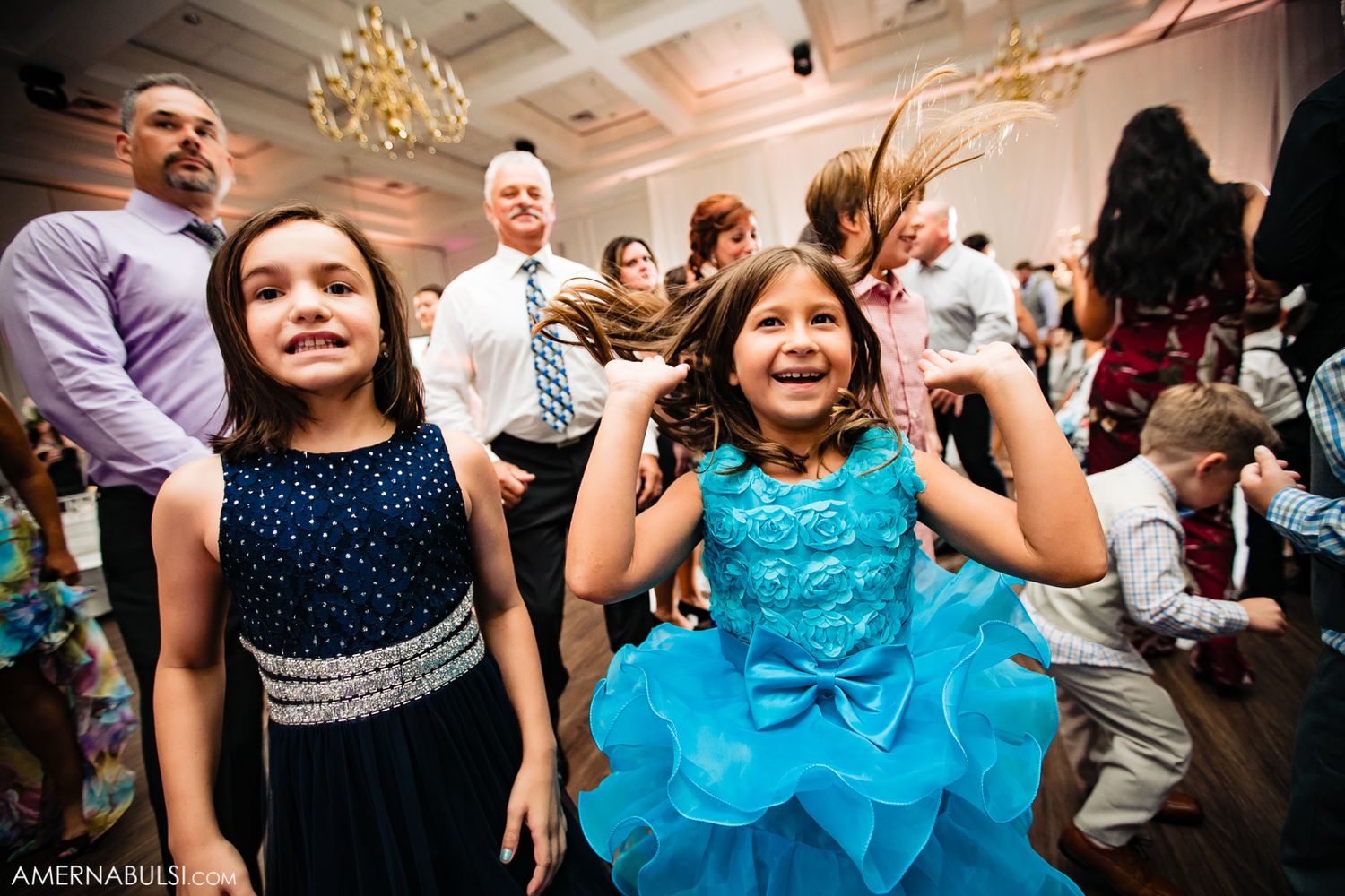 People dancing and celebrating at an indoor event with chandeliers and festive atmosphere.