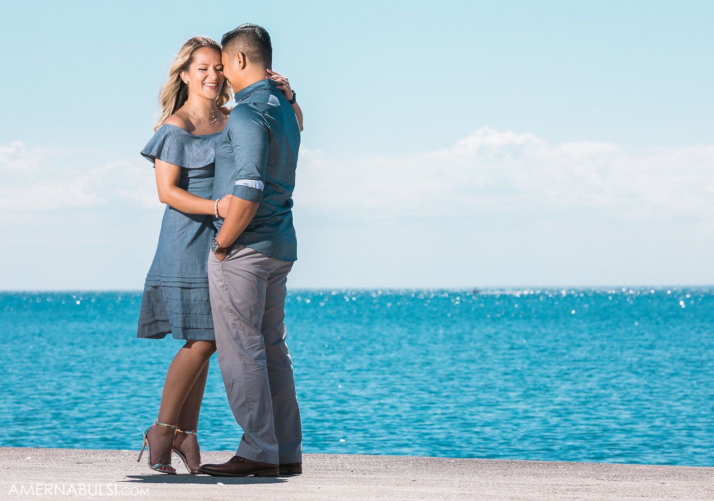 A romantic couple poses together on a beach with turquoise water in the background.