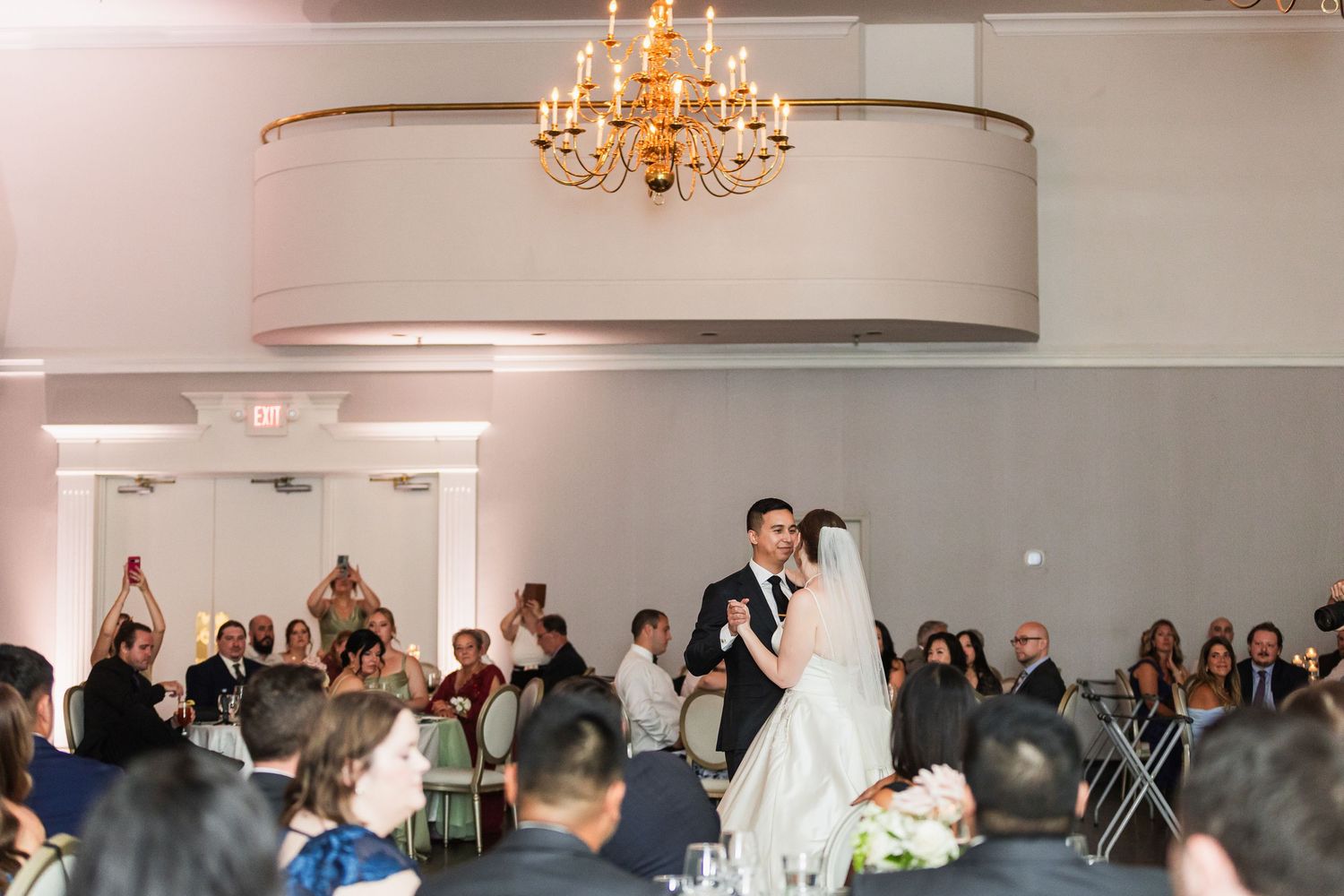 Bride and groom share first dance at elegant wedding reception with chandelier and guests watching.
