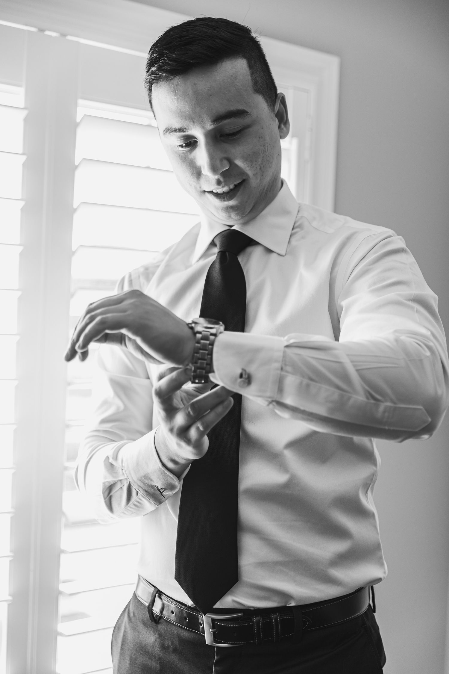 Groom in white dress shirt and black tie fastens his watch cufflinks while smiling in black and white photo.