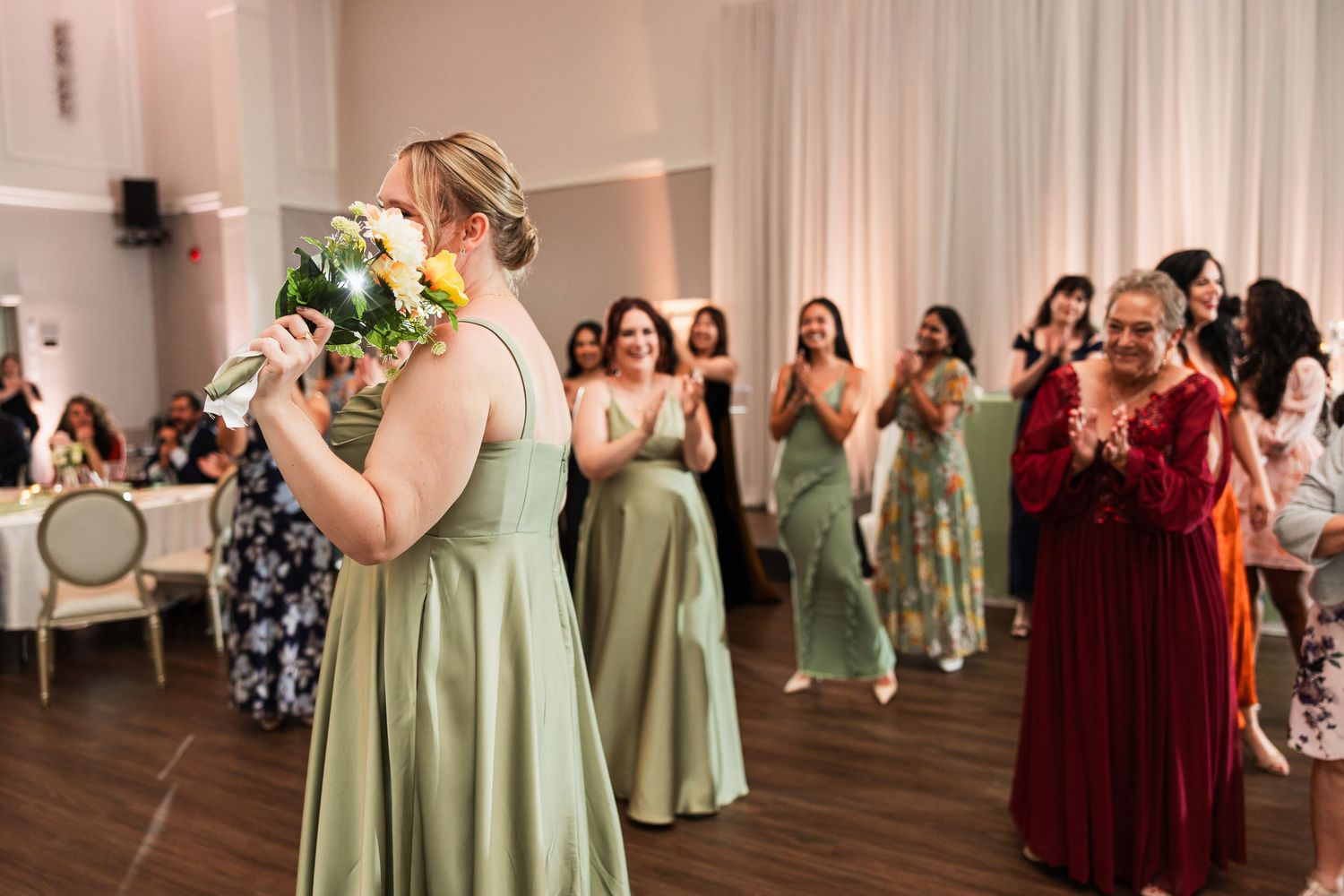 Bridesmaid in sage green gown prepares to toss bridal bouquet to excited guests at elegant wedding reception.