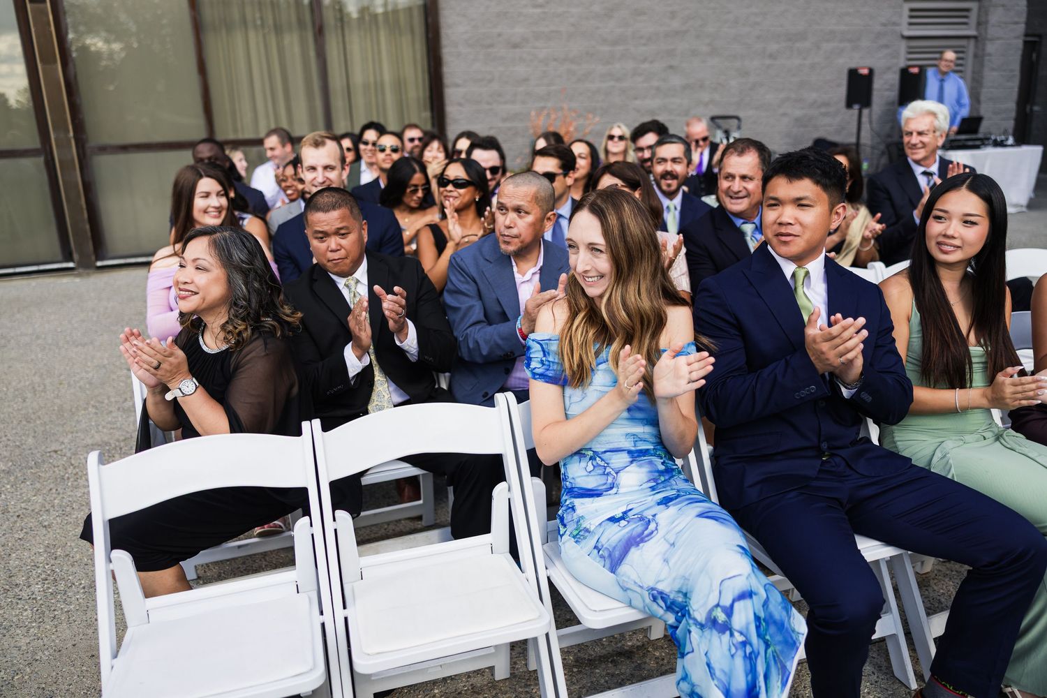 Guests clapping and smiling while seated in white chairs at an outdoor wedding ceremony.