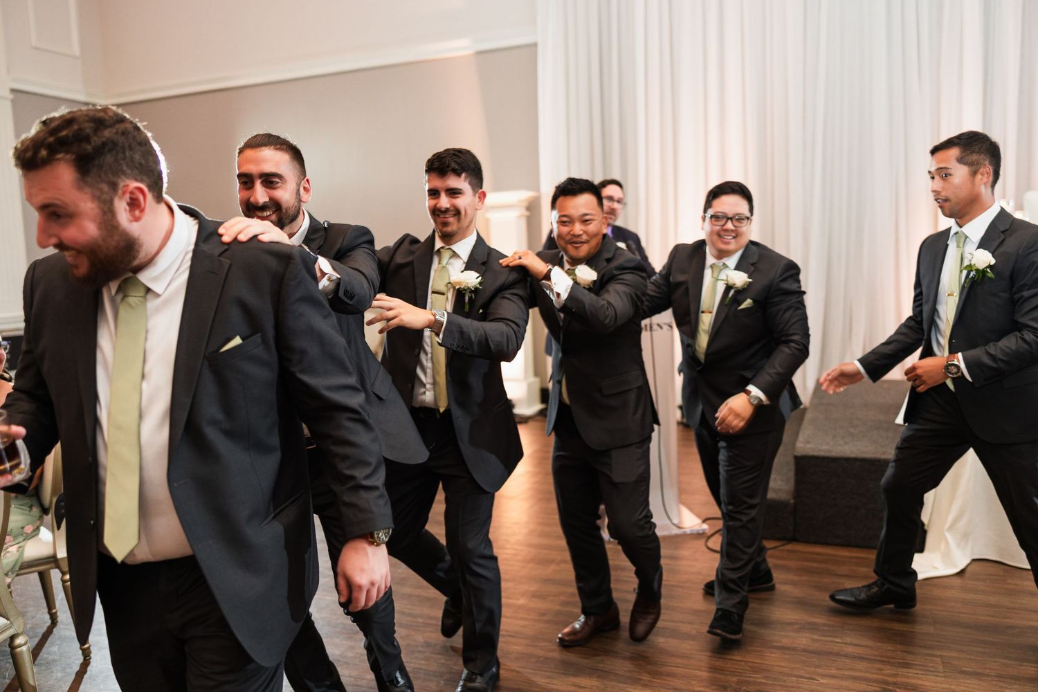 Groomsmen in dark suits with light ties dancing and celebrating joyfully at a wedding reception.