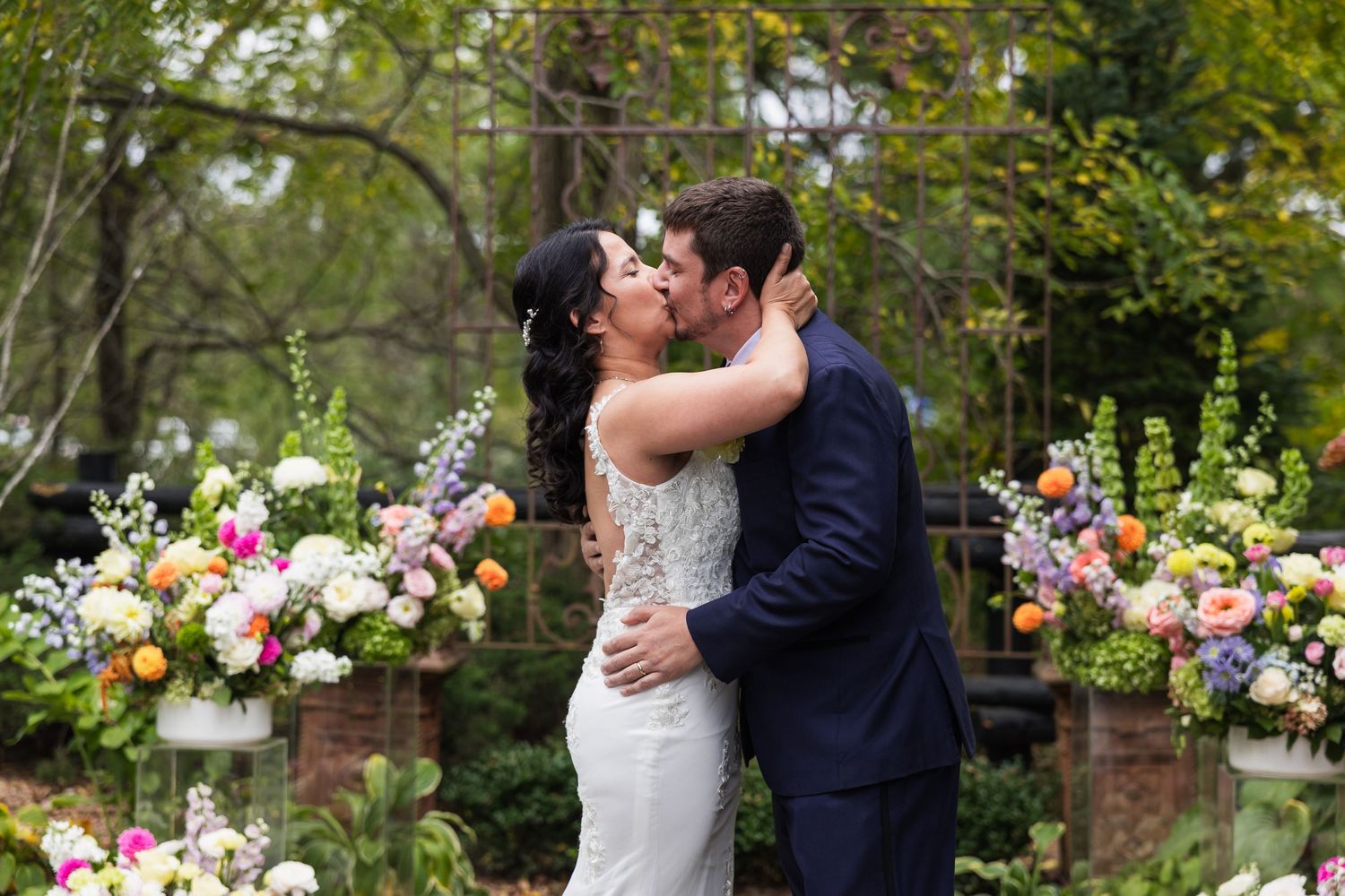 Bride and groom share first kiss at outdoor wedding ceremony surrounded by colorful floral arrangements.