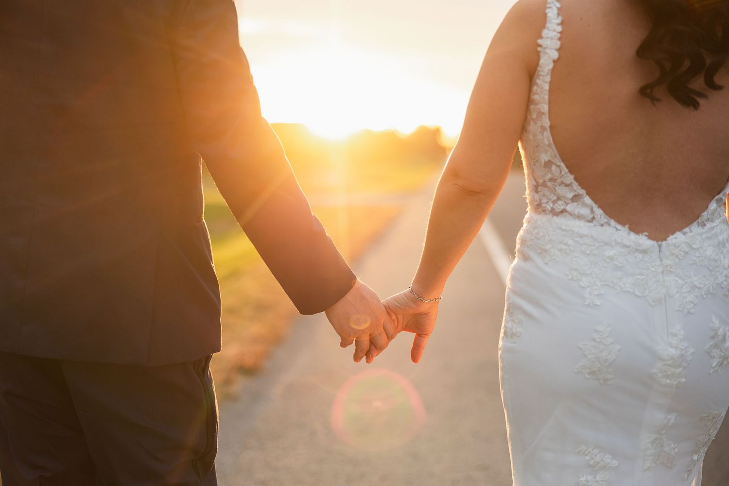 Bride and groom holding hands at sunset on a country road, bride wearing a lace backless wedding gown.
